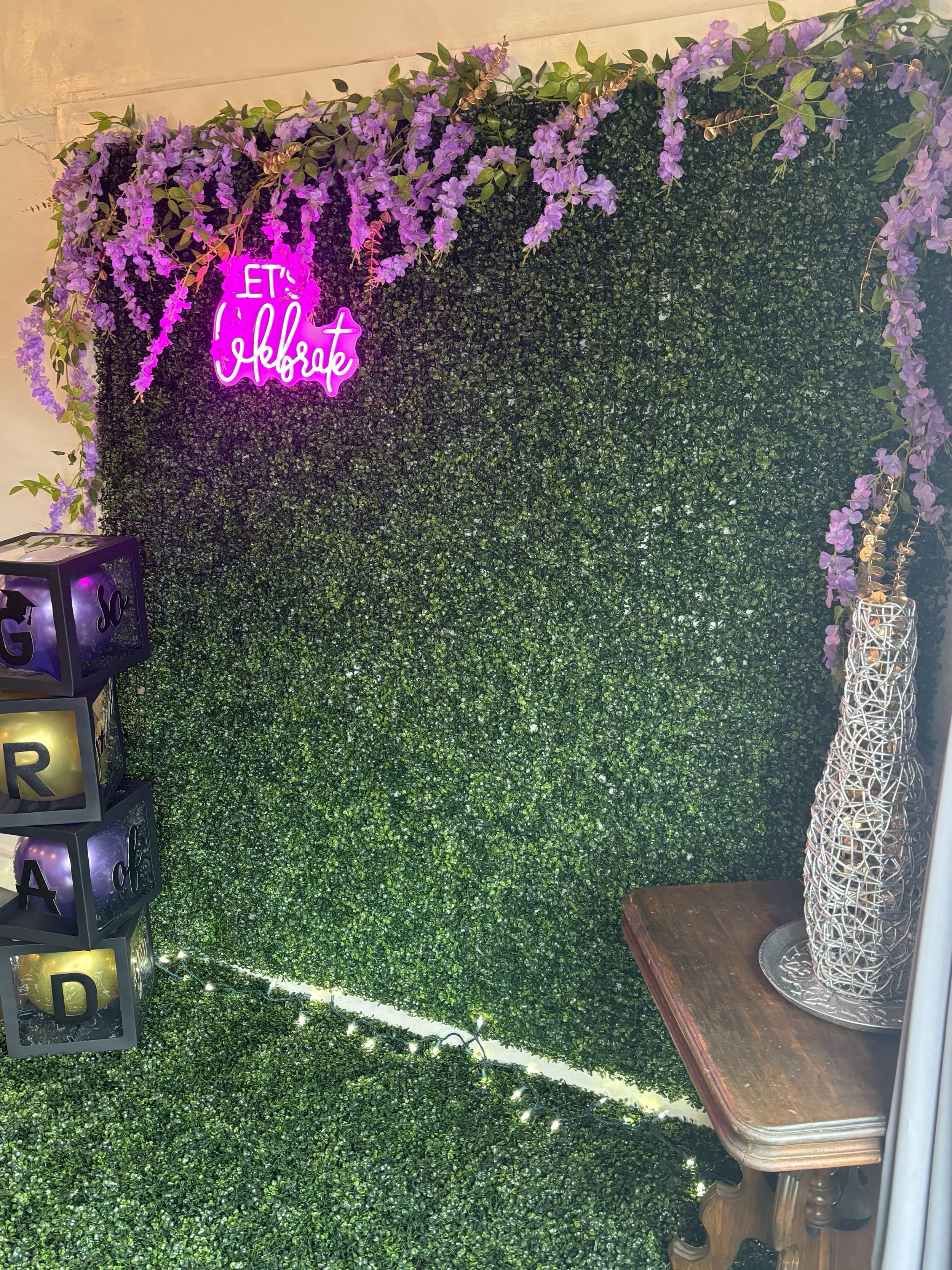 Decorative wall with lush green foliage, purple flowers at the top, a bright pink neon sign that reads 'Let's Celebrate', illuminated alphabet blocks spelling 'GRA'D' on the left, a wooden table with decorative woven vases on the right, and white string lights along the floor.