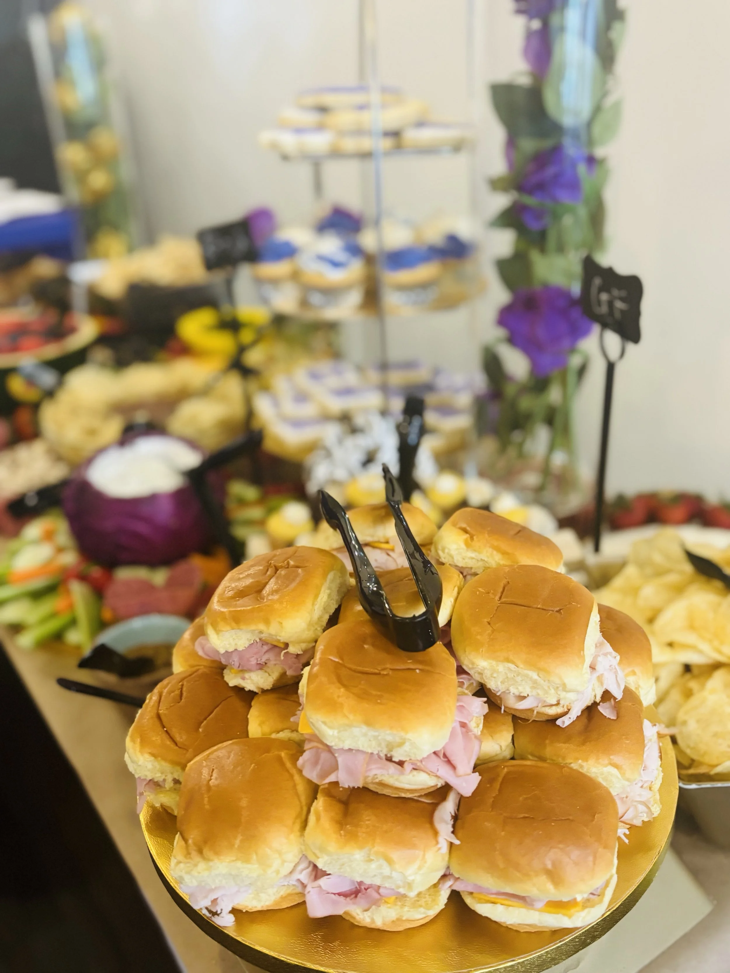 A tray of small sandwiches with ham and cheese arranged in a pyramid. In the background, a buffet of various foods, including salads, chips, and desserts, with purple flowers and chalkboard labels.