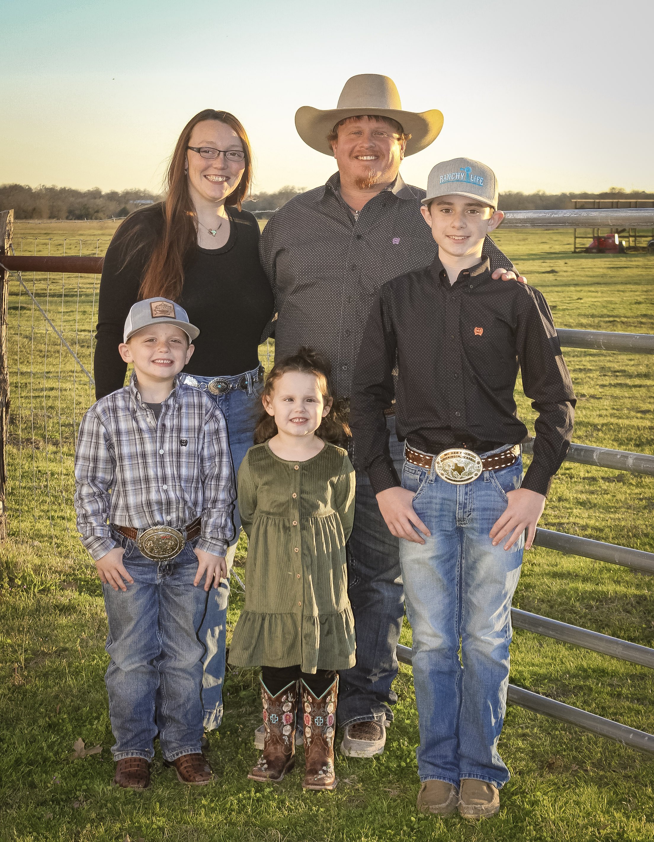 Family of five dressed in cowboy attire standing outside on a farm during sunset, with a fence and open field in the background.