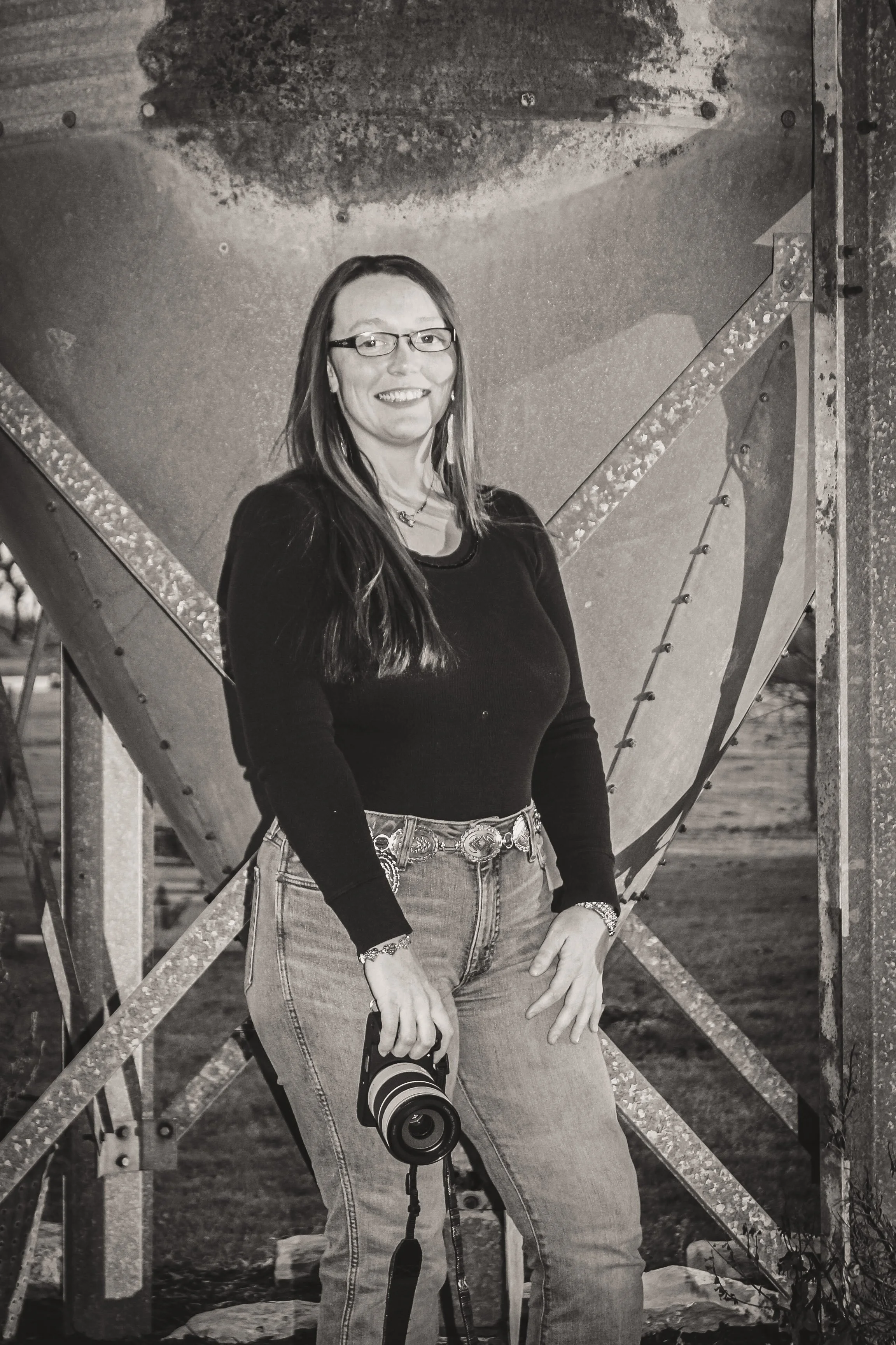 A smiling woman standing outdoors at duck in front of a large metal structure, holding a camera with a long lens.
