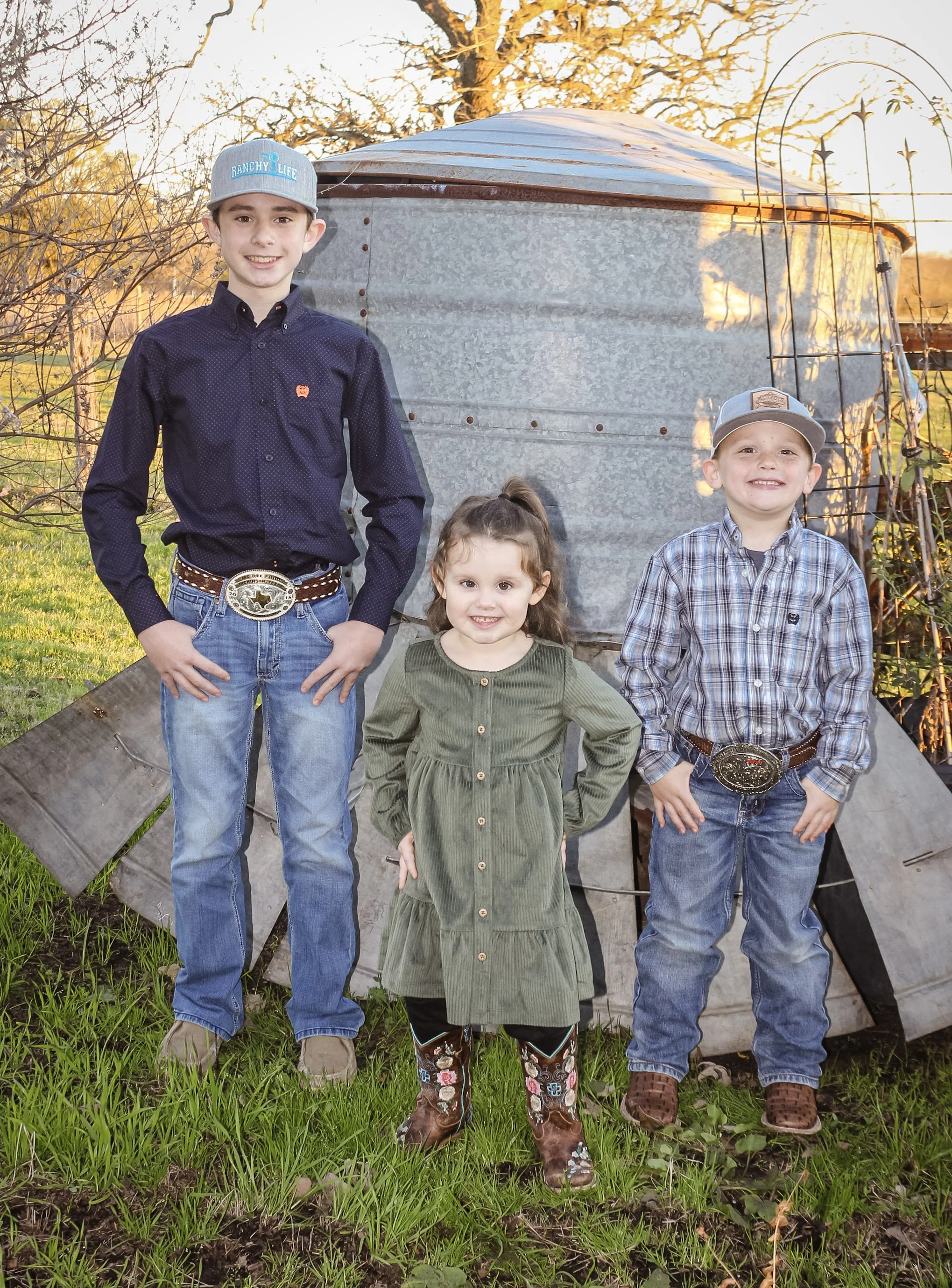Three children, two boys and one girl, standing outdoors on grass in front of a weathered metal structure, likely a small shed or barn, during sunset. The boy on the left wears a gray cowboy hat, a dark button-up shirt, jeans, and western belt buckle. The girl in the middle sports an olive green dress, black leggings, and floral cowboy boots. The boy on the right is dressed in a plaid shirt, jeans, a cowboy hat, and a large belt buckle.