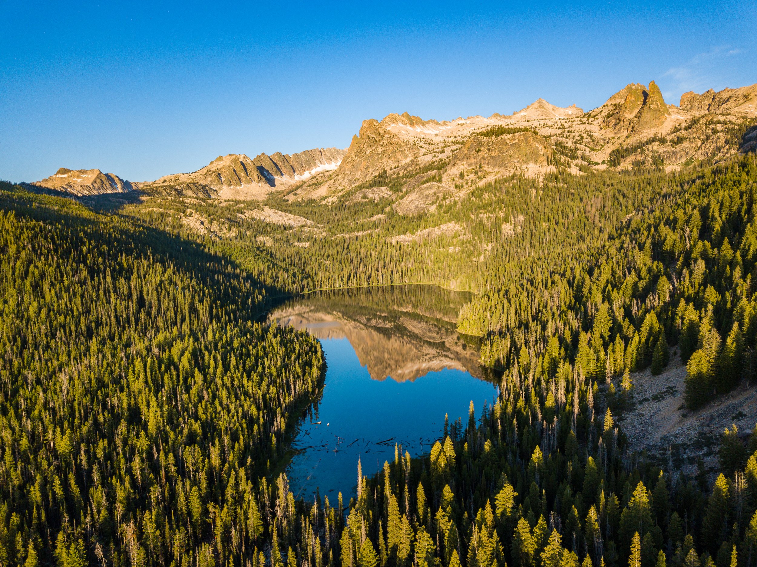 Aerial drone photo of Hell Roaring Lake in the Sawtooth Mountains of Idaho, surrounded by forest and rugged granite peaks.