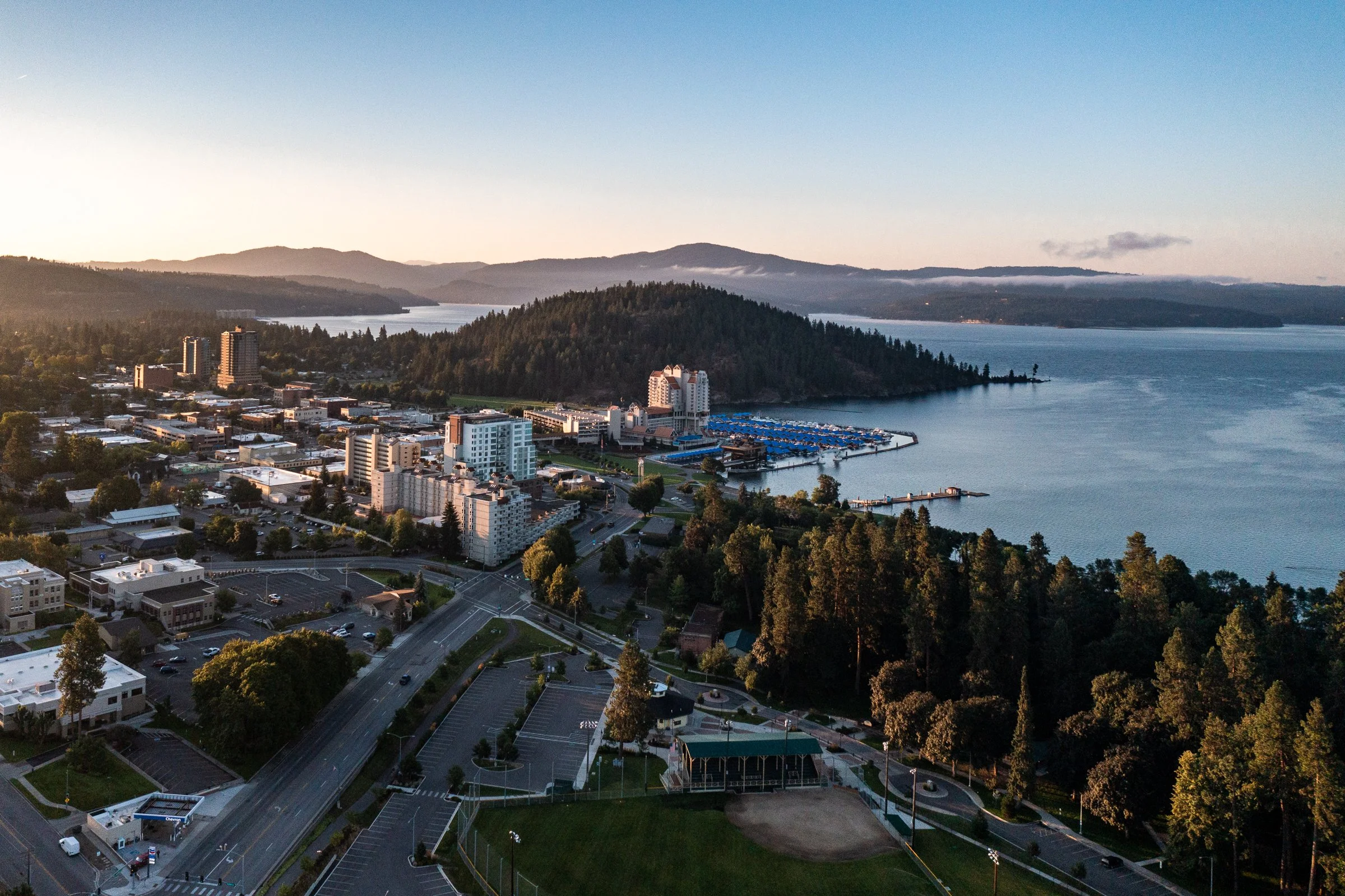 Morning aerial drone photo of Coeur d’Alene, Idaho, showing calm lake water, forested shoreline, and surrounding mountains.
