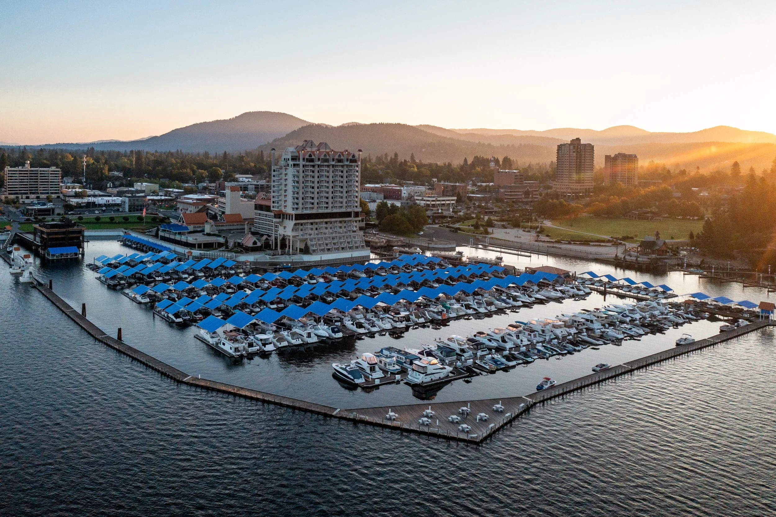 Aerial drone photograph of Coeur d’Alene, Idaho, captured at sunrise by Jon Conti Visuals. This 5K image shows boats docked along the waterfront hotel marina with calm lake reflections and soft morning light over the surrounding mountains — a cinemat