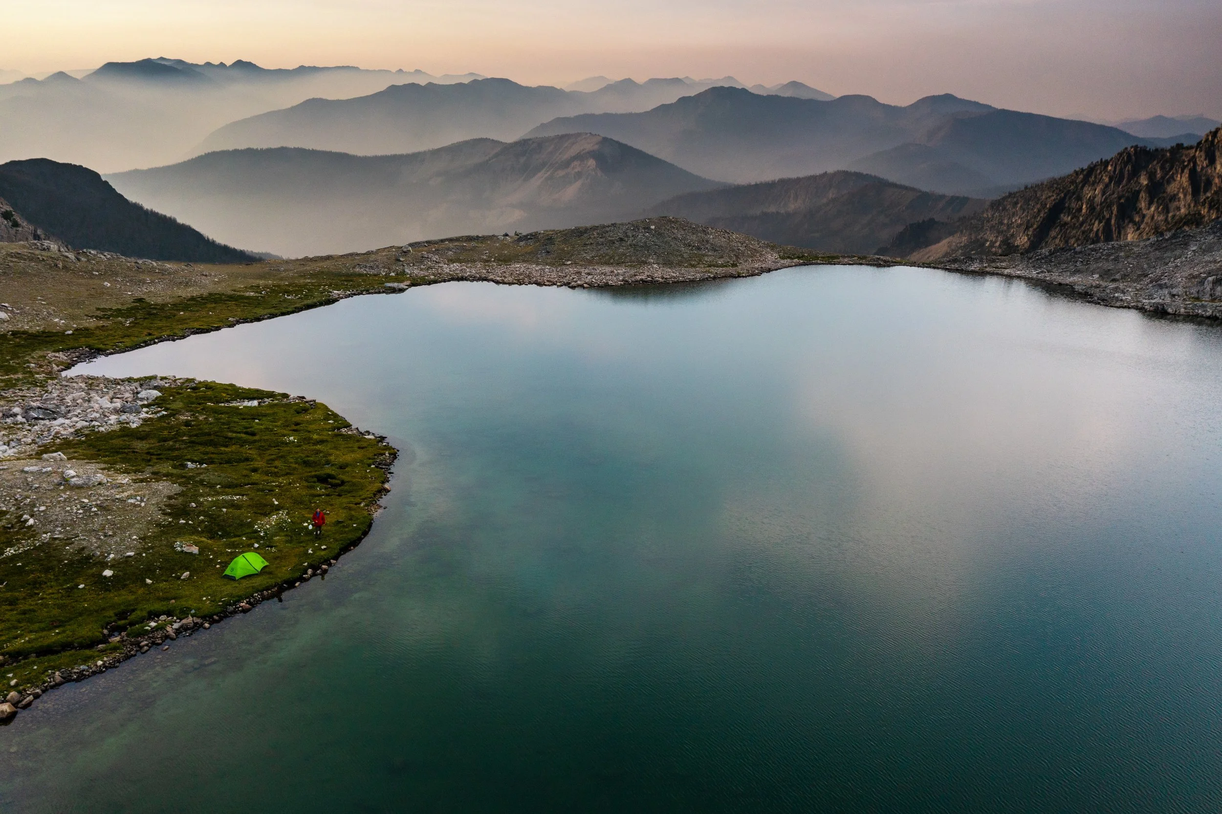Aerial drone photograph of Goat Lake in the Pioneer Mountains near Sun Valley, Idaho, captured by Jon Conti Visuals. This 5K image shows the deep blue alpine water surrounded by rocky peaks and early summer snowfields — a cinematic view of Idaho’s hi