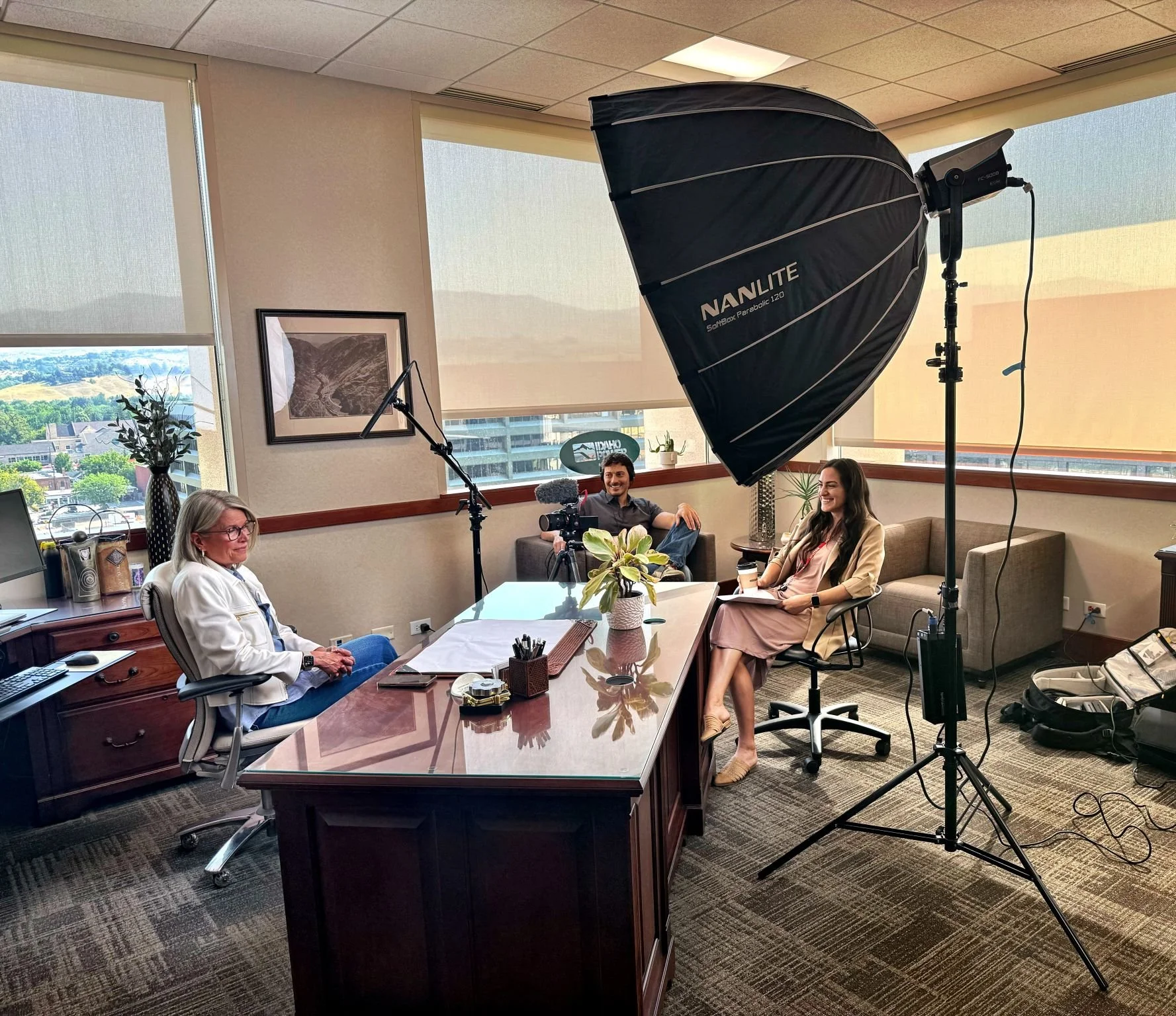 A woman sits at a desk during a video recording setup with studio lighting, a camera, and two people sitting on a couch in a bright office with large windows and mountains visible outside.
