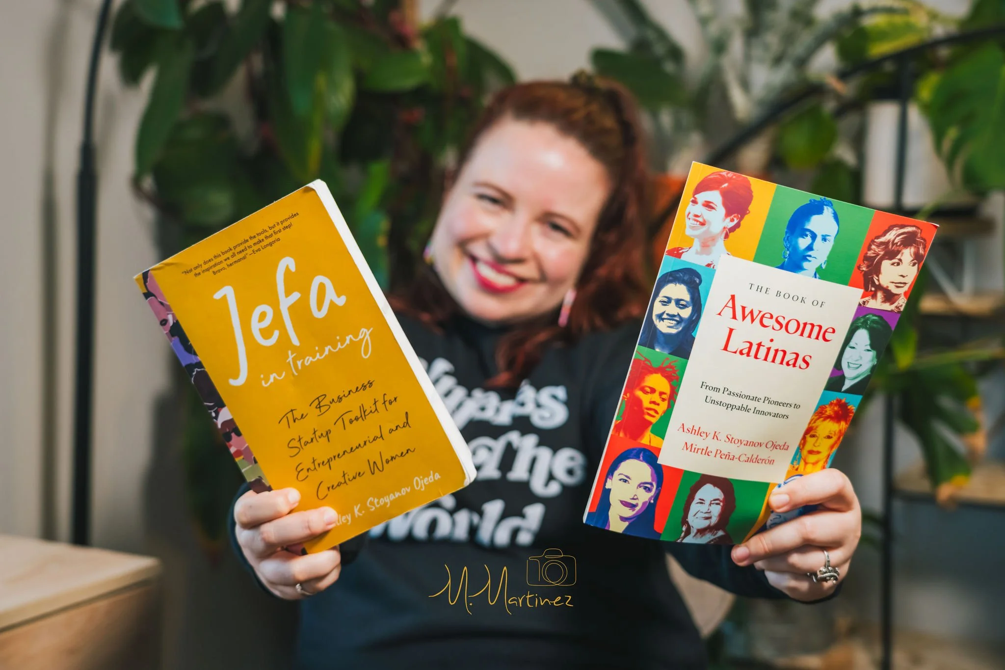 A woman with reddish hair smiling as she holds two colorful books, one titled 'Leta in training' and the other 'The Book of Awesome Latinas,' with a green plant in the background.