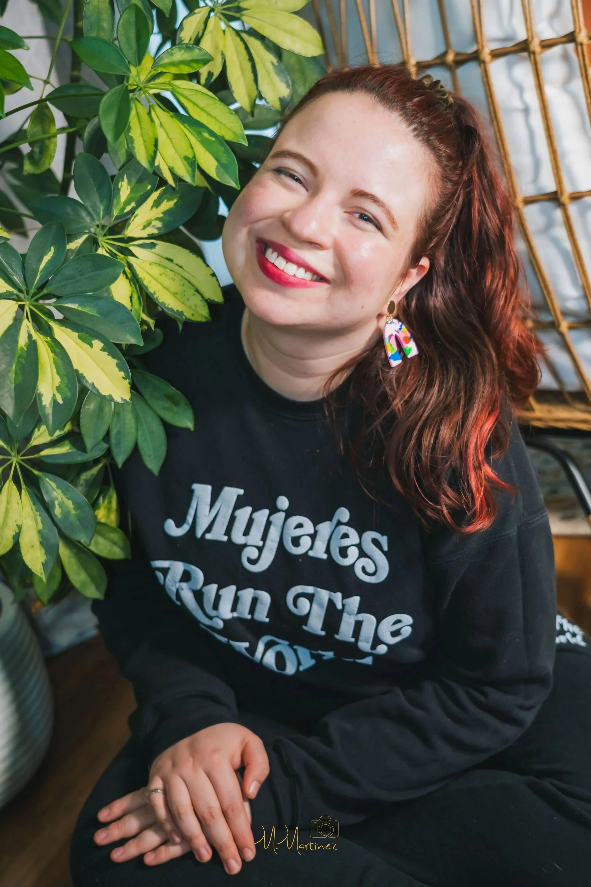 A woman with red hair and colorful earrings, smiling, is sitting beside a large green and yellow variegated plant. She is wearing a black shirt with white text. The background includes bamboo-style furniture and a white quilted fabric.