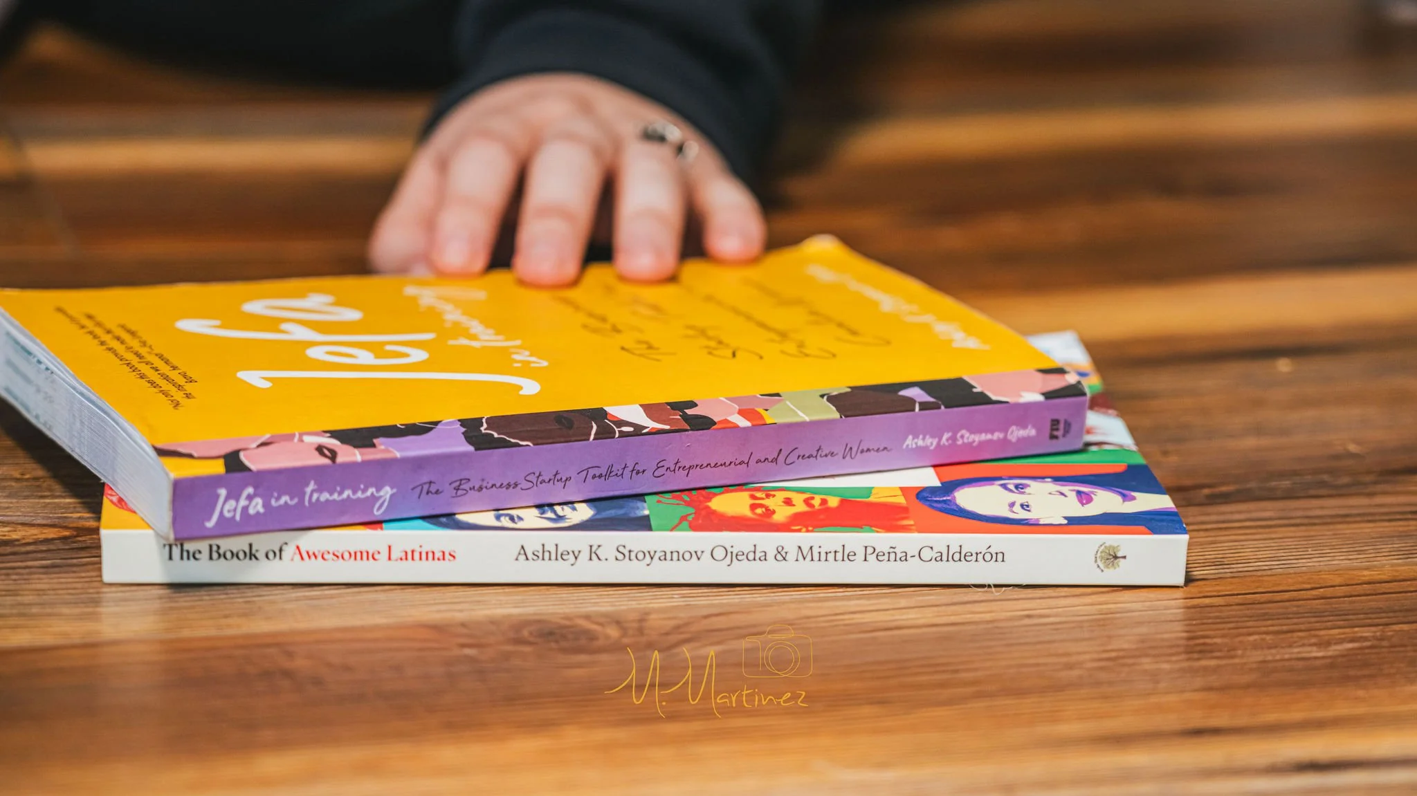 A hand resting on top of three books stacked on a wooden surface. The books are titled 'Jefa in Training', 'The Book of Awesome Latinas', and another book with colorful illustrations on the cover.