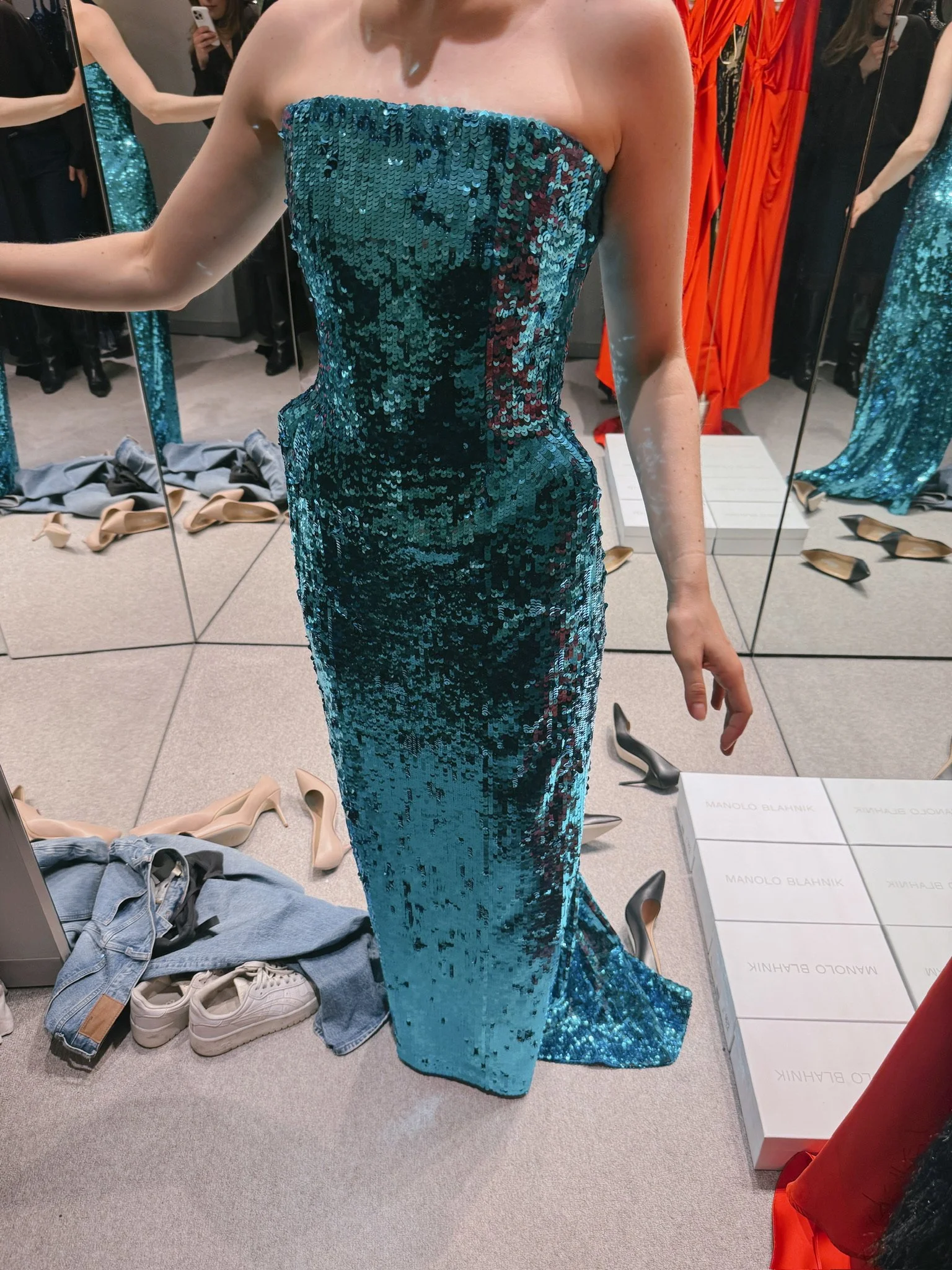 A woman is trying on a strapless, floor-length, blue sequined dress in a fitting room. The room has mirrors, various shoes, and clothing items on the floor.