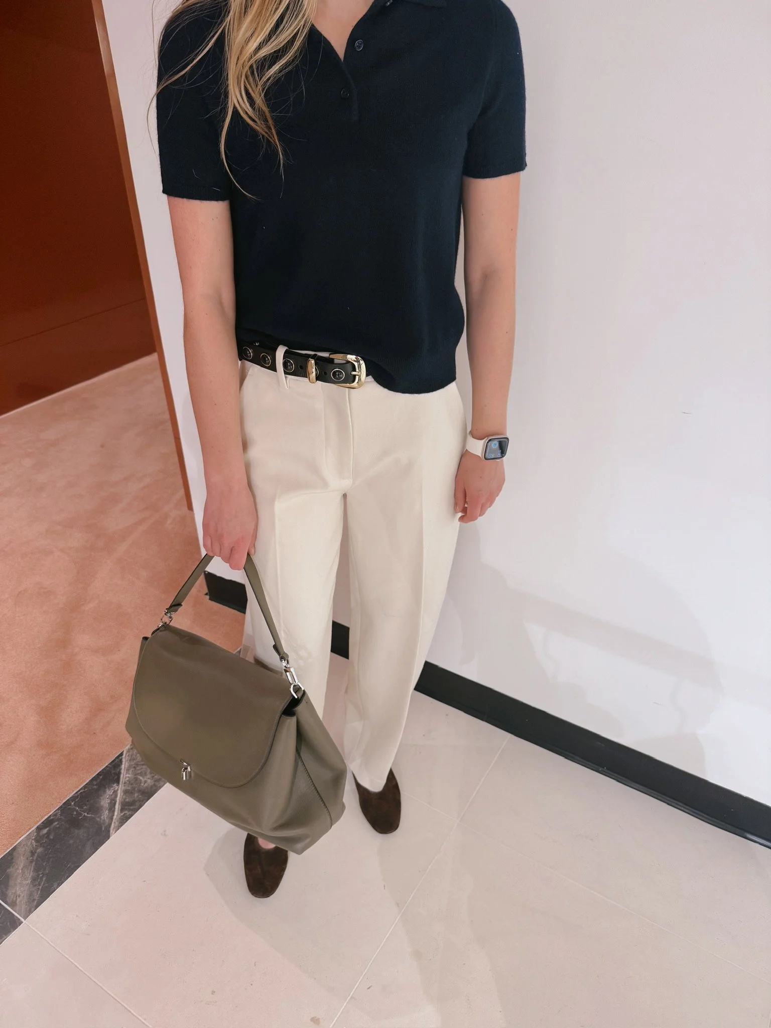 A person wearing a black polo shirt, cream-colored pants, brown shoes, holding a beige handbag, and wearing a smartwatch.