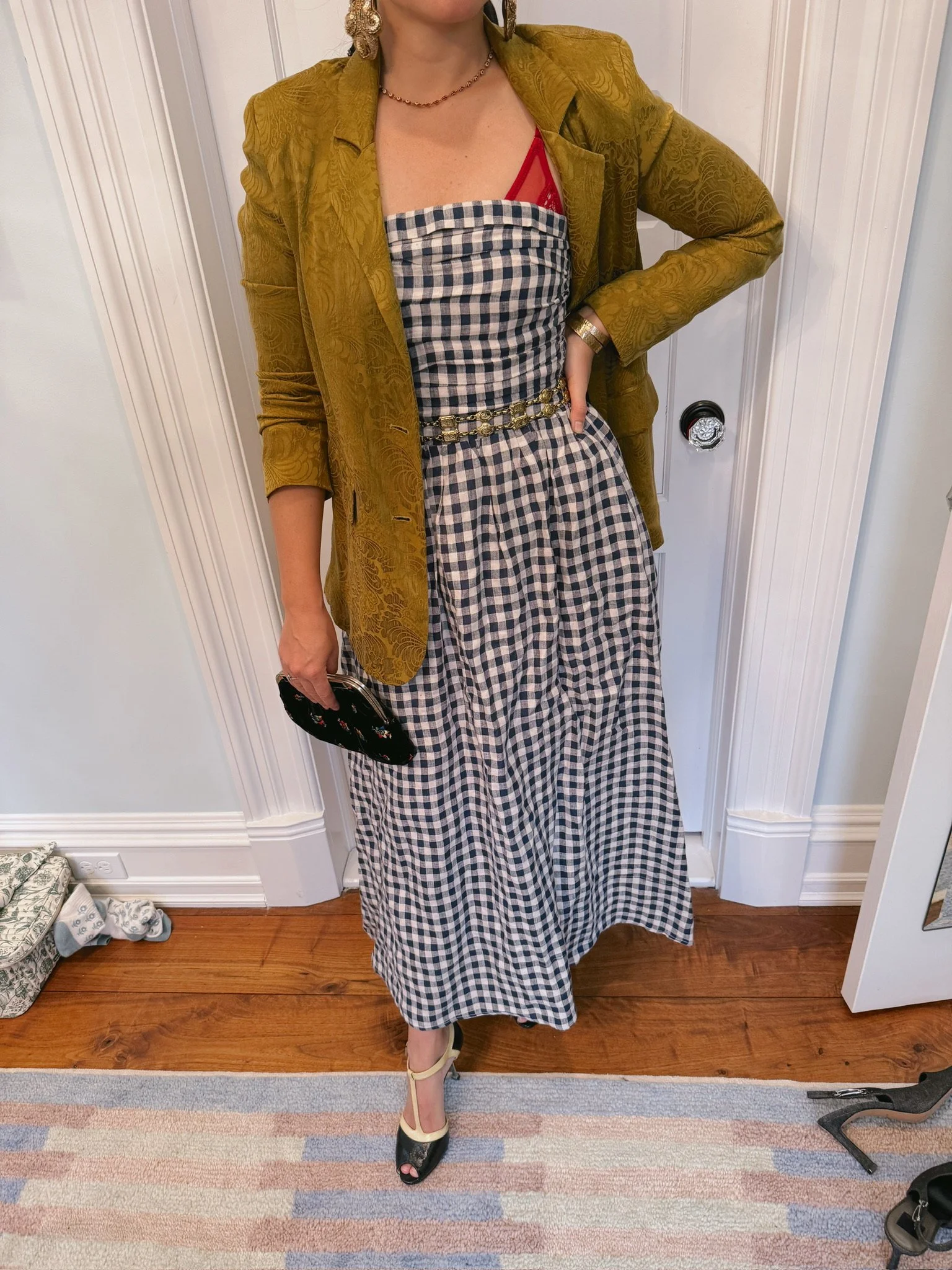 Woman wearing a yellowish-green blazer over a black and white checkered strapless dress with a red bra visible, holding a black clutch, wearing high heels, in front of a white door and wall with hardwood flooring and a rug.