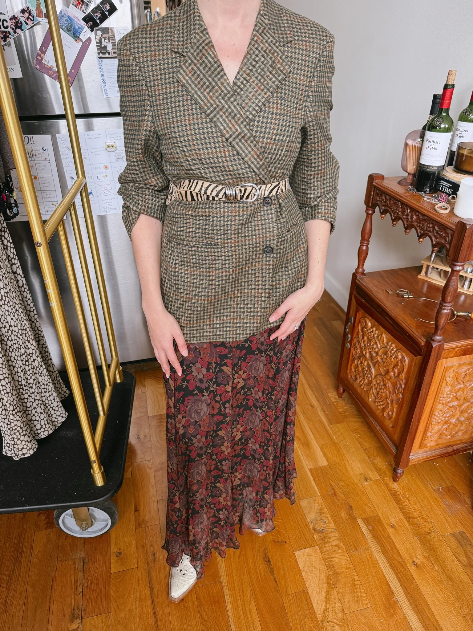 Person wearing a brown and green plaid blazer and matching skirt, with a zebra-print belt, white shoes, standing in a room with a wooden floor, a small wooden table, and a refrigerator covered with photos and notes.