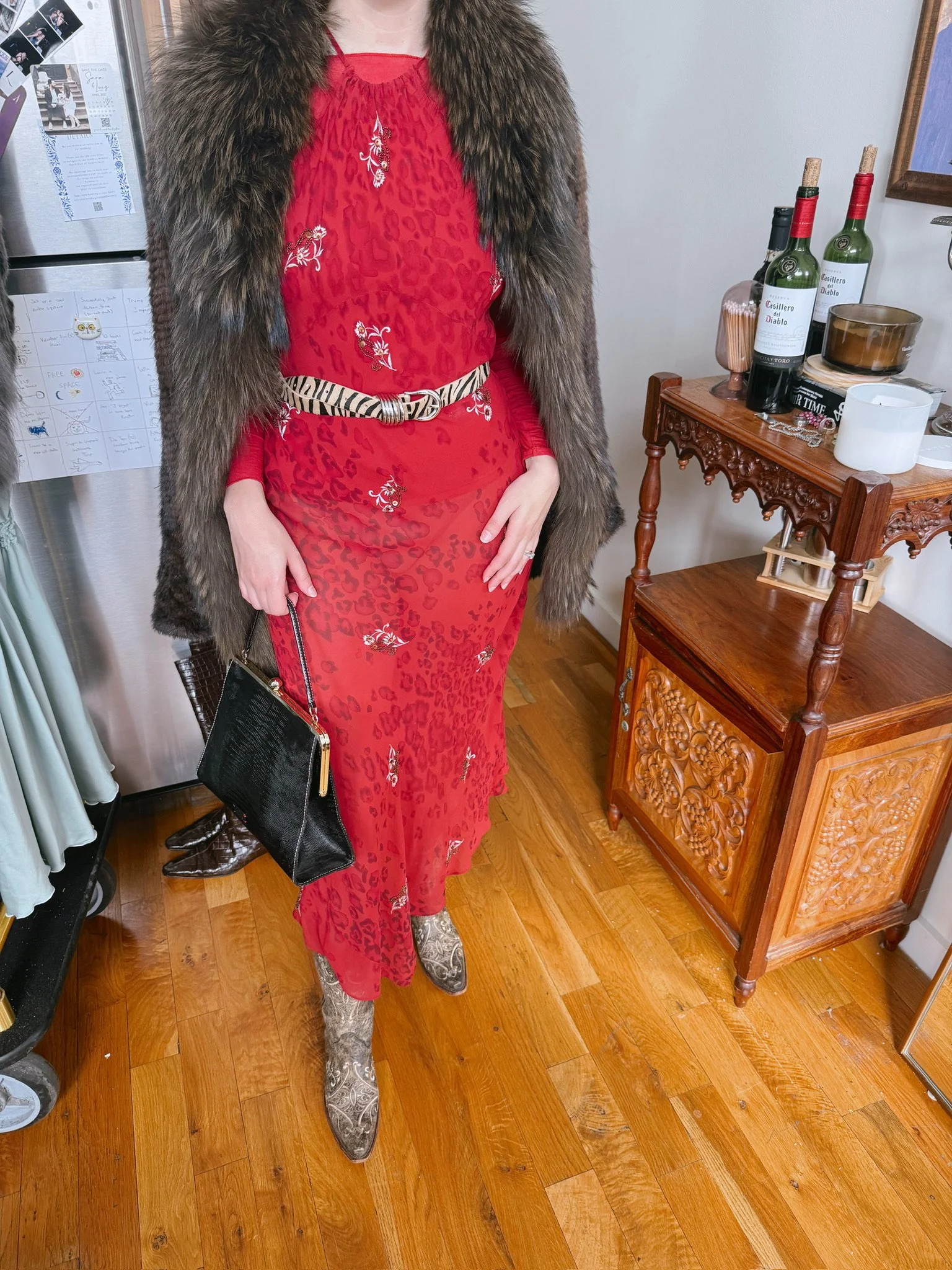 Person wearing a red dress with white floral embroidery, a leopard print belt, a fur coat, and patterned cowboy boots, holding a black purse, standing indoors near a wooden side table with bottles of wine and various decor items.