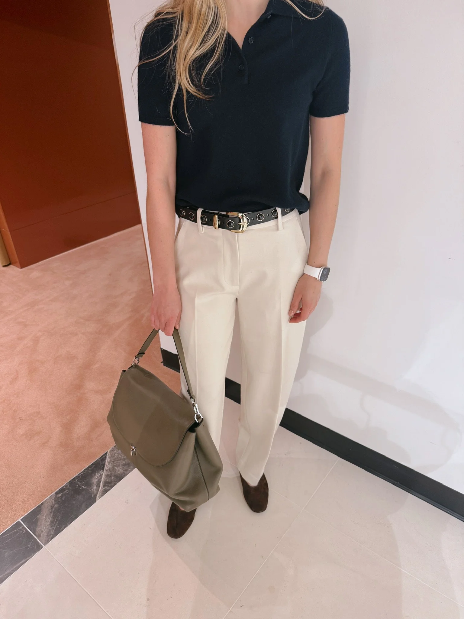 A woman dressed in a navy polo shirt, beige trousers, and brown shoes, holding a gray handbag, standing indoors against a white wall.