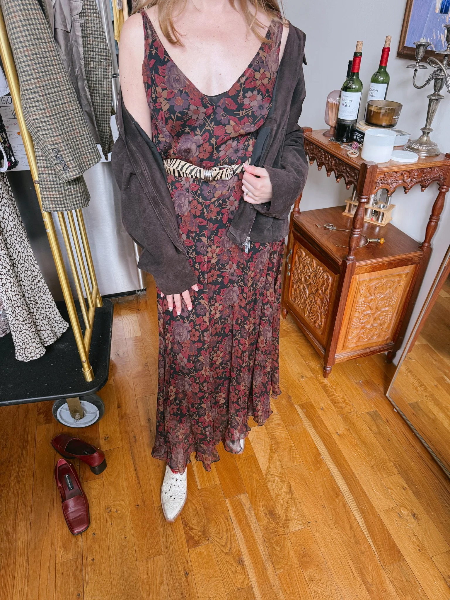A person taking a mirror selfie wearing a floral maxi dress with a dark jacket draped over their shoulders. They are wearing white shoes with cut-out details and standing on wooden flooring. To their left, there are red shoes on the floor. Behind them, there is a clothing rack with jackets and a wooden cabinet with bottles and decorative items.