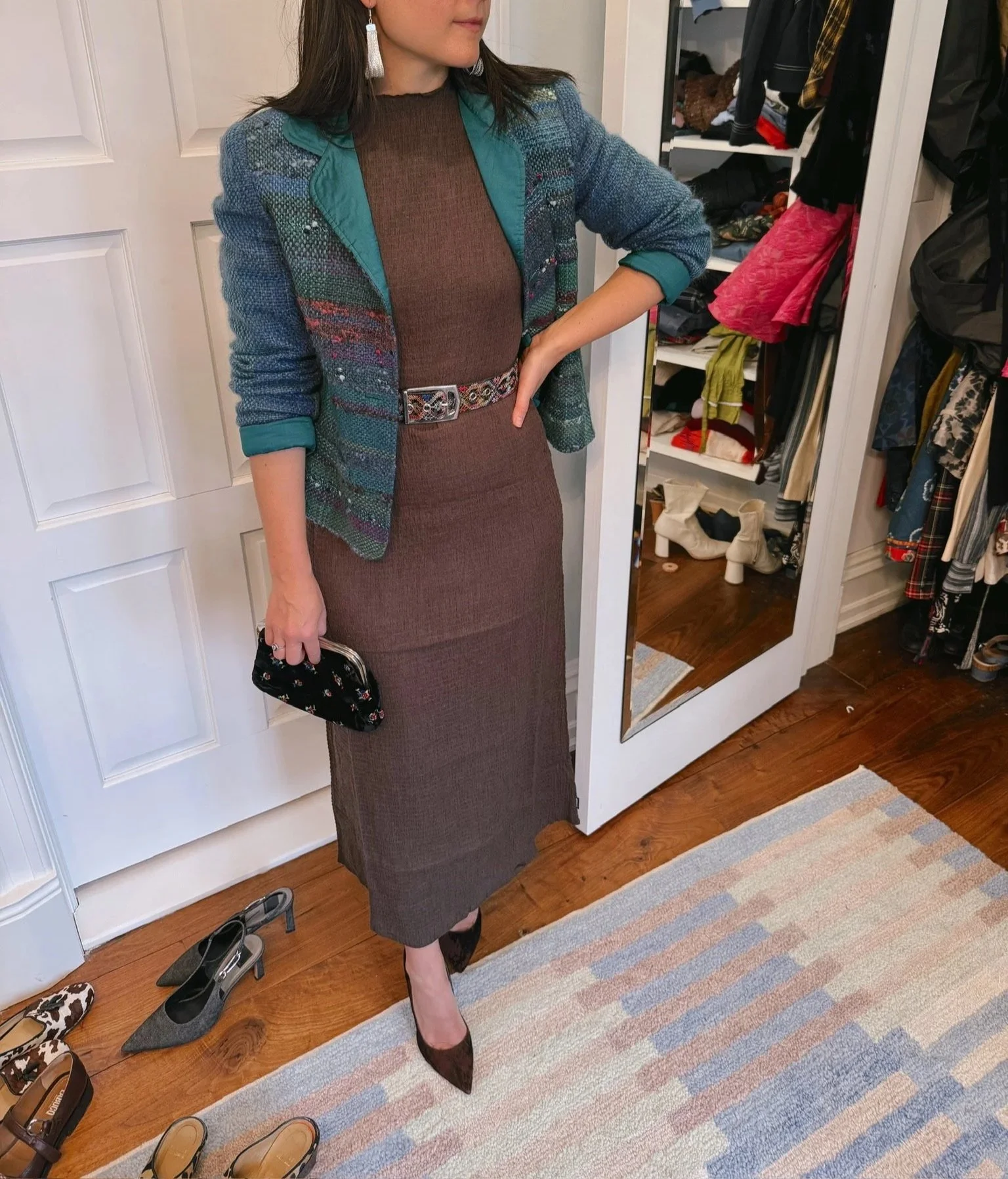 A woman dressed in a brown dress with a multicolored plaid blazer, high heels, and tassel earrings, standing in a walk-in closet with shoes and clothing visible.
