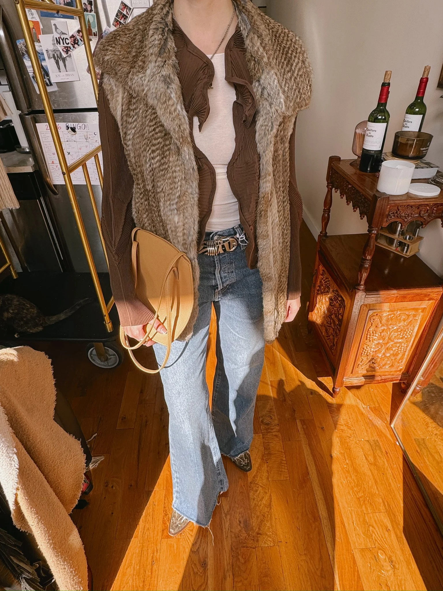 A person wearing a brown outfit with a mix of textures, including a fur and patterned coat, a white top, and wide-legged jeans, holding a tan purse. The setting appears to be a cozy indoor space with wooden furniture and a fridge decorated with photos and notes.