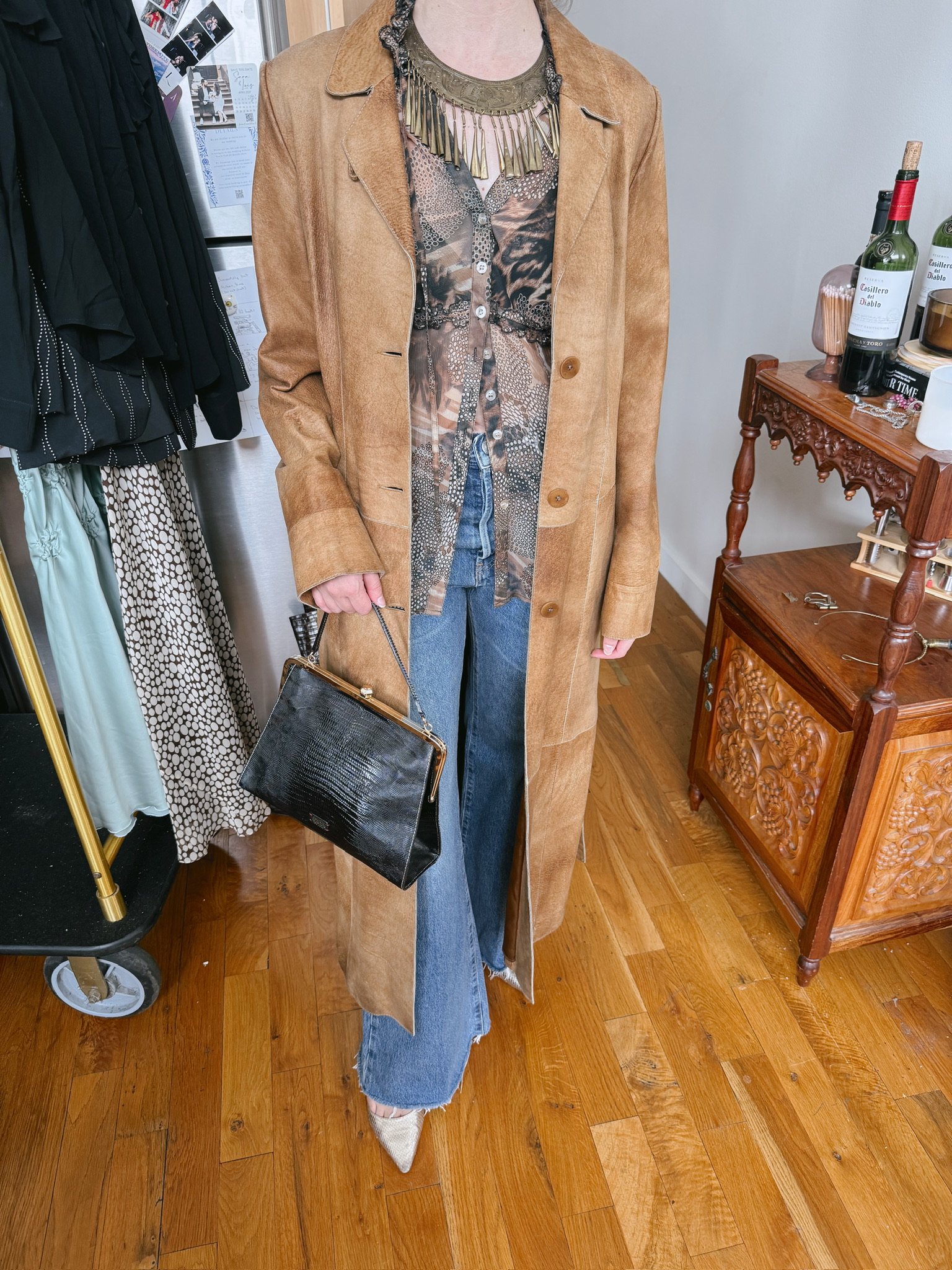 Person in a tan long coat, patterned blouse, and jeans holding a black handbag with gold hardware, standing on a wooden floor near a wooden cabinet with bottles and decor.