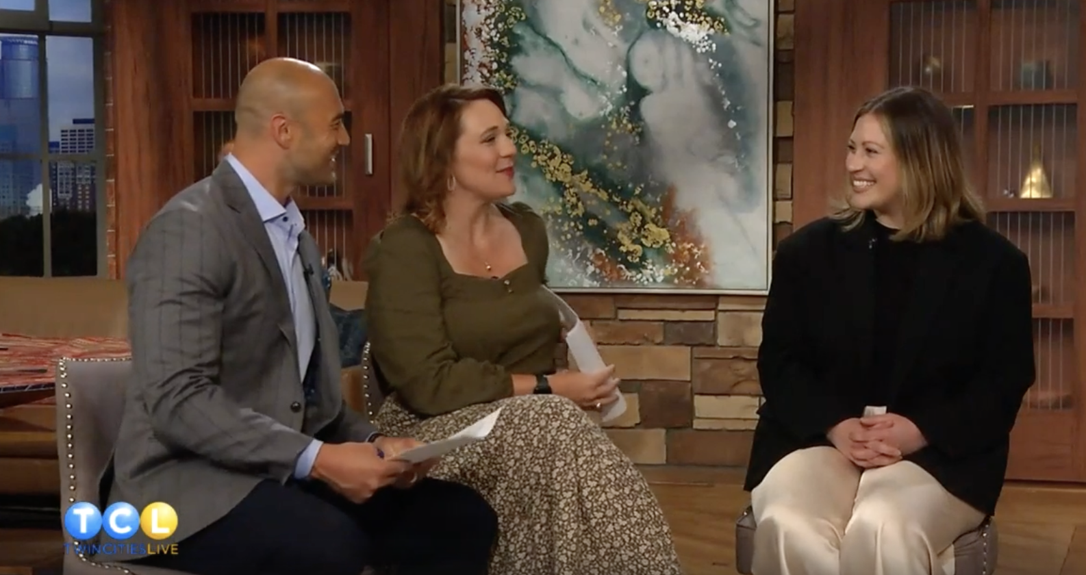 Three people sitting and talking in a studio. The man on the left wears a gray suit, the woman in the middle has brown hair and wears a green blouse, and the woman on the right has blonde hair and wears black and beige. There is a large abstract painting and wooden cabinets behind them, with cityscape visible through the window.