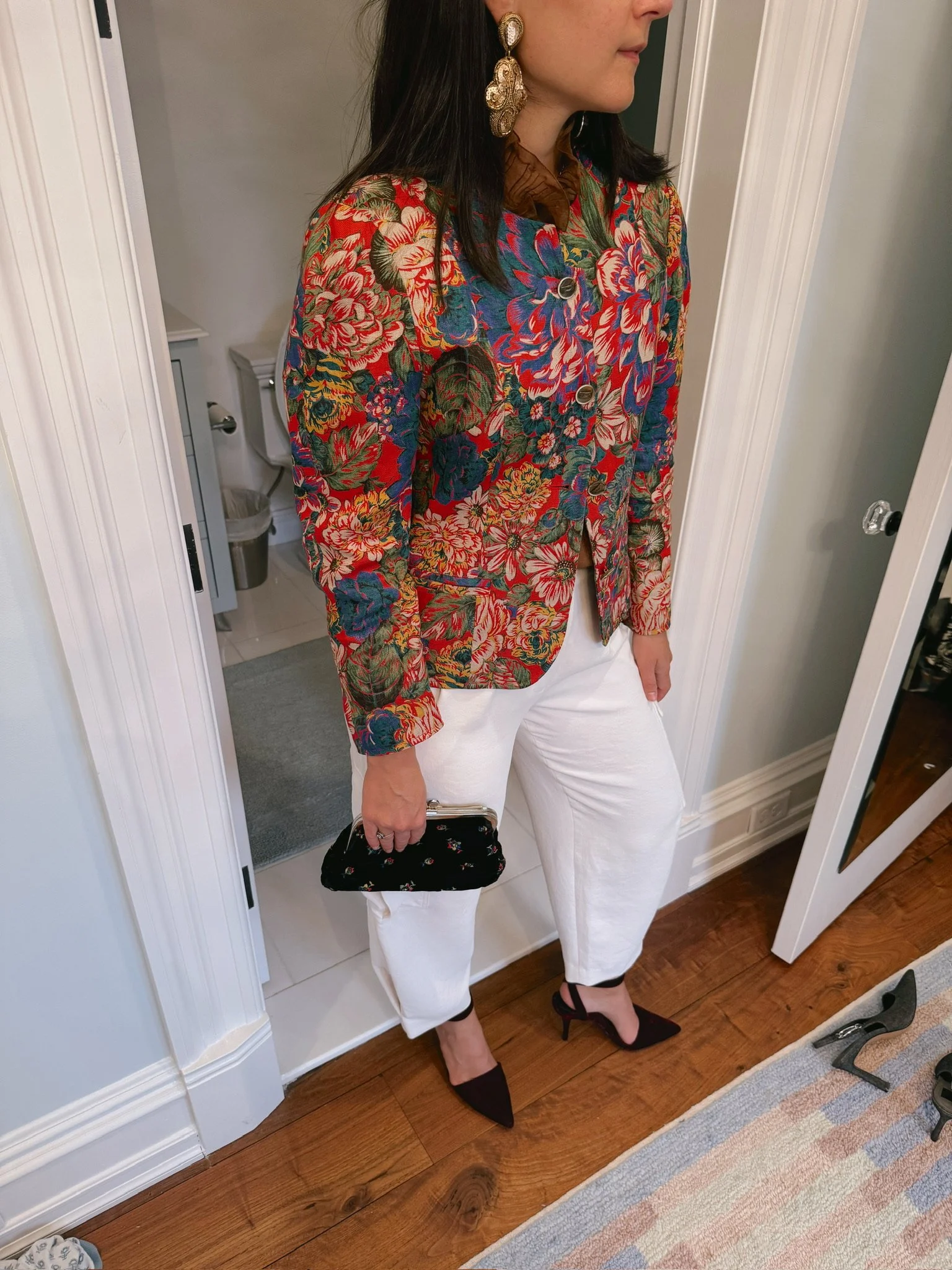 Woman dressed in a floral patterned jacket, white pants, black high heels, holding a black purse with floral design, large gold earrings, standing indoors near a bathroom door.