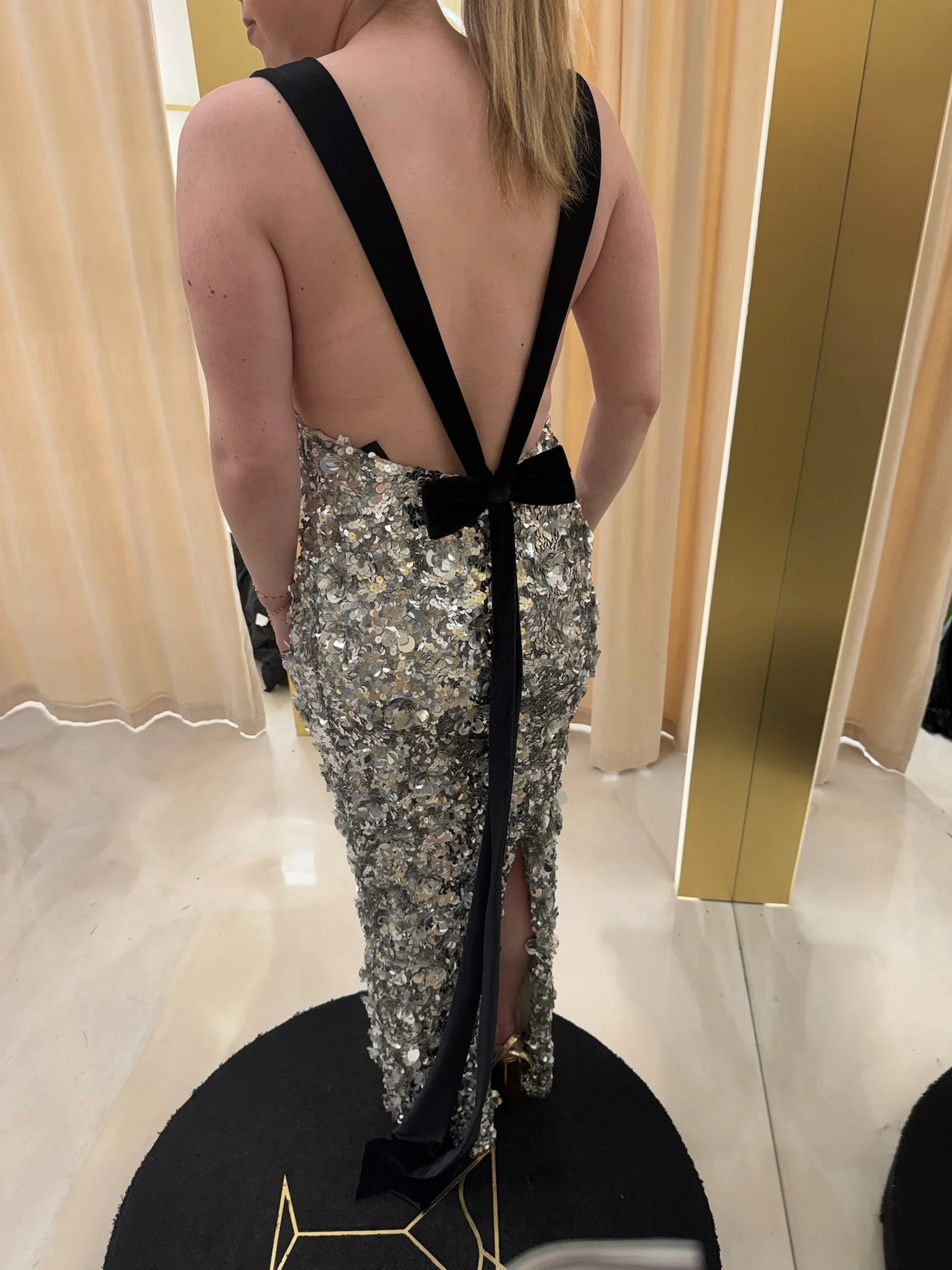 Back view of a woman in a silver sequin dress with a low open back, black straps, and a bow at the waist, standing in front of yellow curtains.
