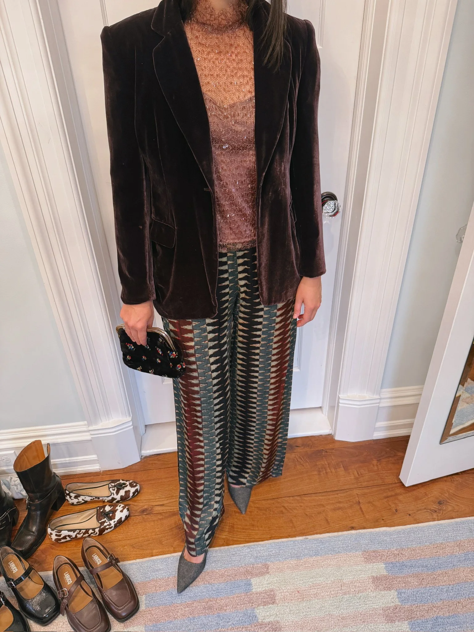 Woman wearing a dark blazer, glittery top, patterned wide-leg pants, gray high heels, holding a small black handbag with floral embroidery.