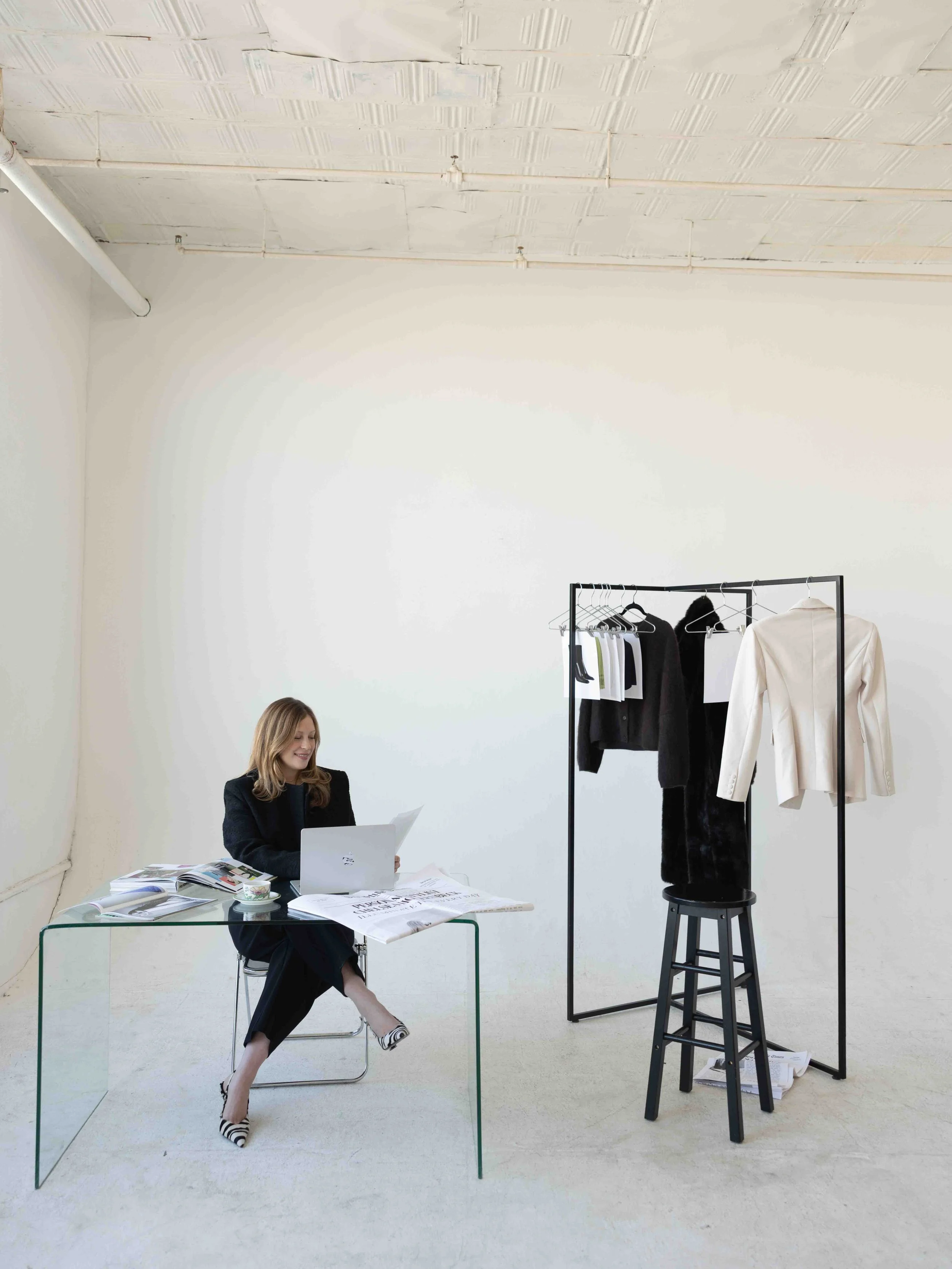 Personal stylist Chelsea Hollenbeck working at a glass desk beside a clothing rack in a bright fashion studio.