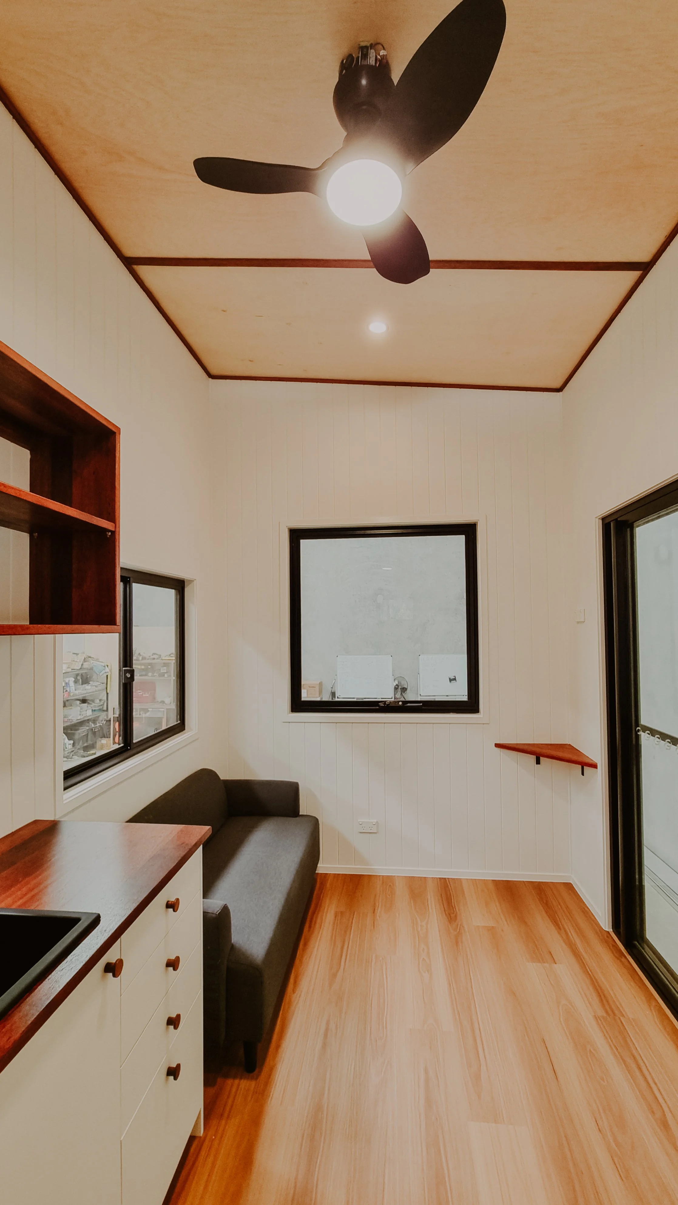 A small room with white walls, wooden flooring, a black sofa, a window, and a ceiling fan with three blades and a light.