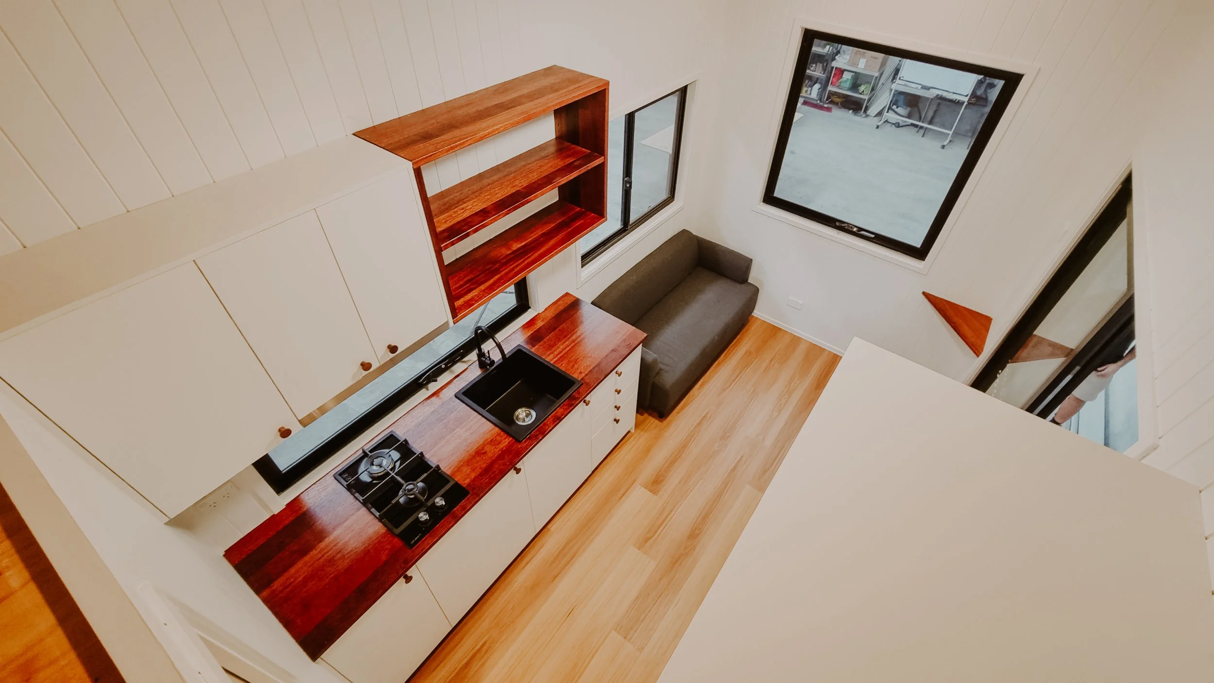 A small kitchen with white cabinets, a black sink, a gas stove, wooden countertops, and an open shelving unit. There is a gray sofa along the wall near two windows and a door leading outside.
