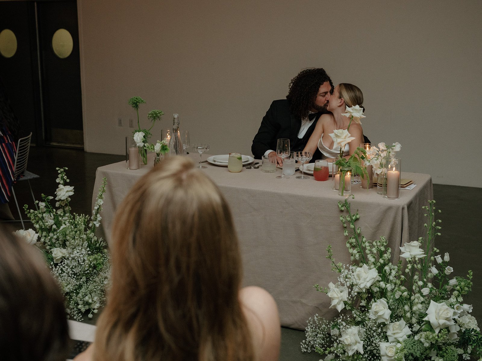  A newlywed couple kissing while sitting at a table surrounded by floral arrangements  