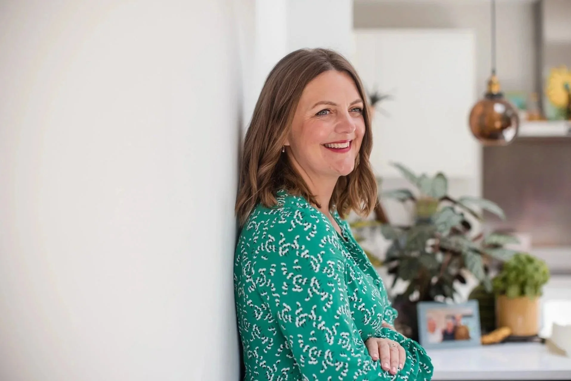 A woman with wavy brown hair smiling and leaning against a white wall, wearing a green blouse with a white and black pattern, in an indoor space with plants and decor in the background.