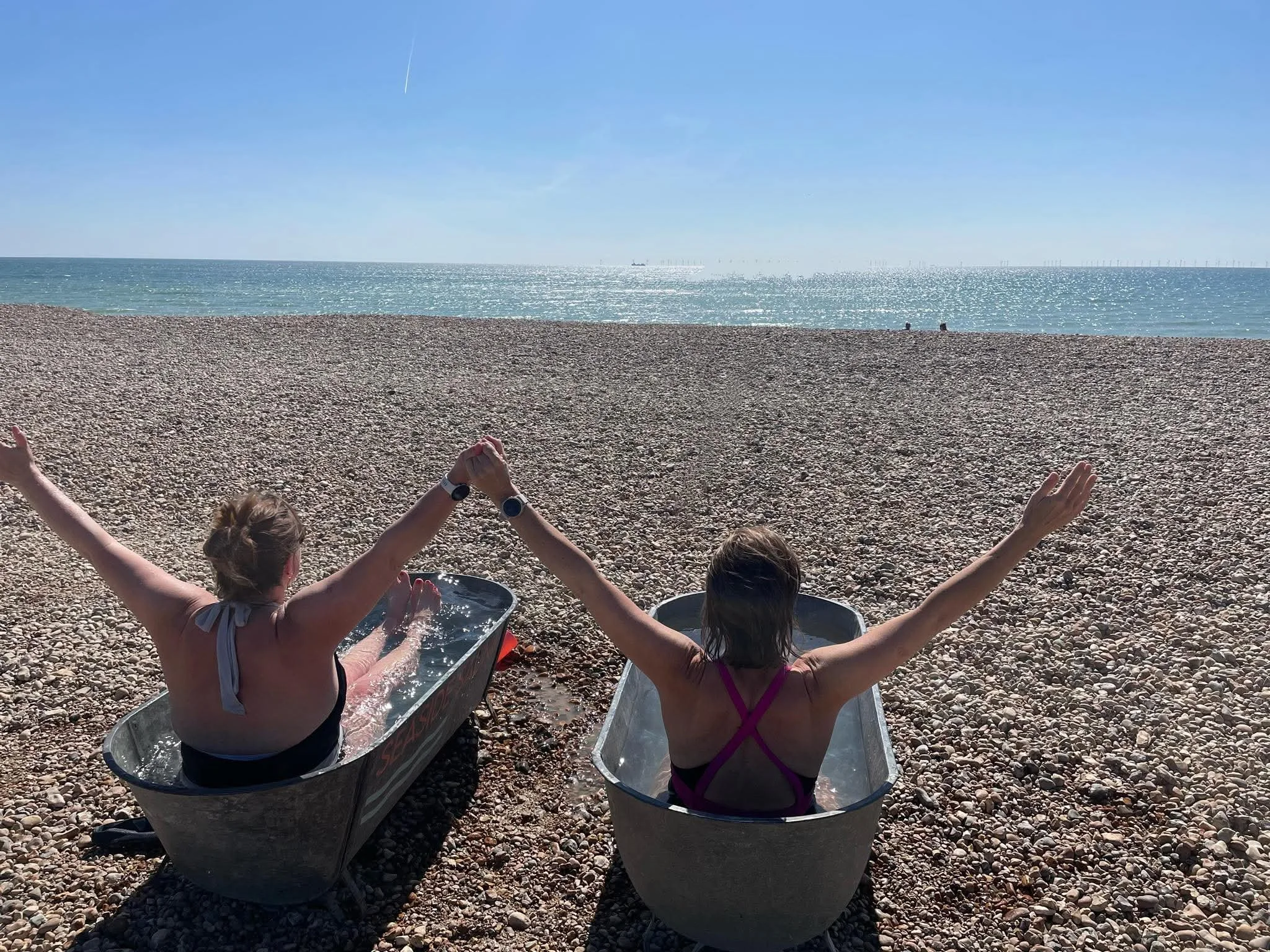 Two adults sitting in metal tubs at the beach with arms raised, facing the ocean, under clear blue sky.
