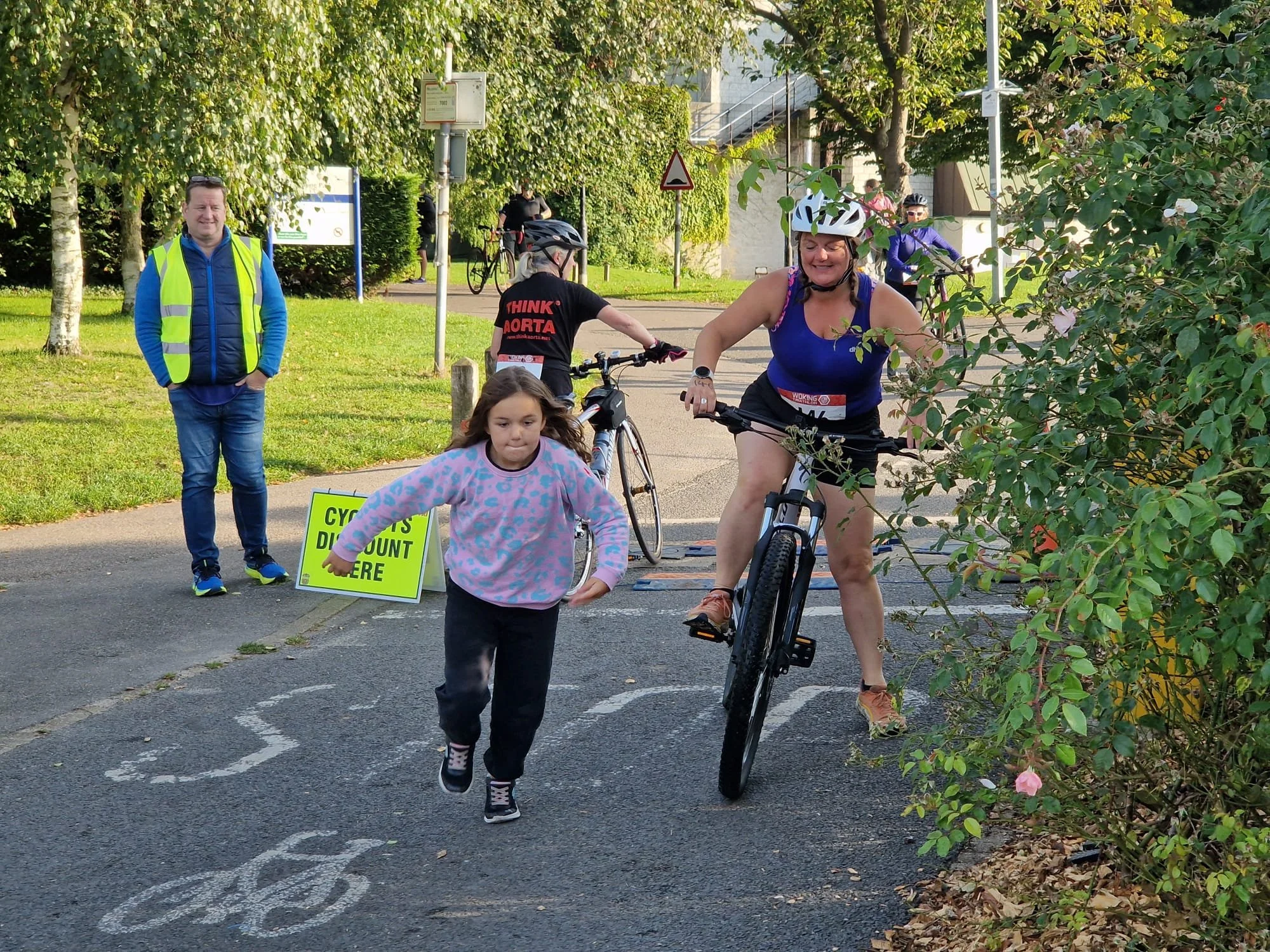 People participating in a cycling event, with a woman riding a bicycle, child walking in front, and several cyclists and a man standing on the side, with trees, signs, and a building in the background.