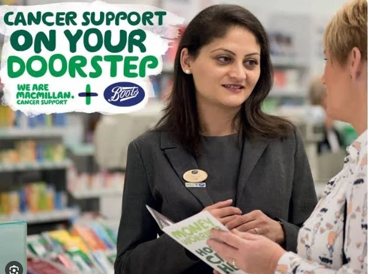 A woman in a store talking to a customer, with a sign behind them promoting cancer support through Macmillan and Boots, and a brochure about money or savings.