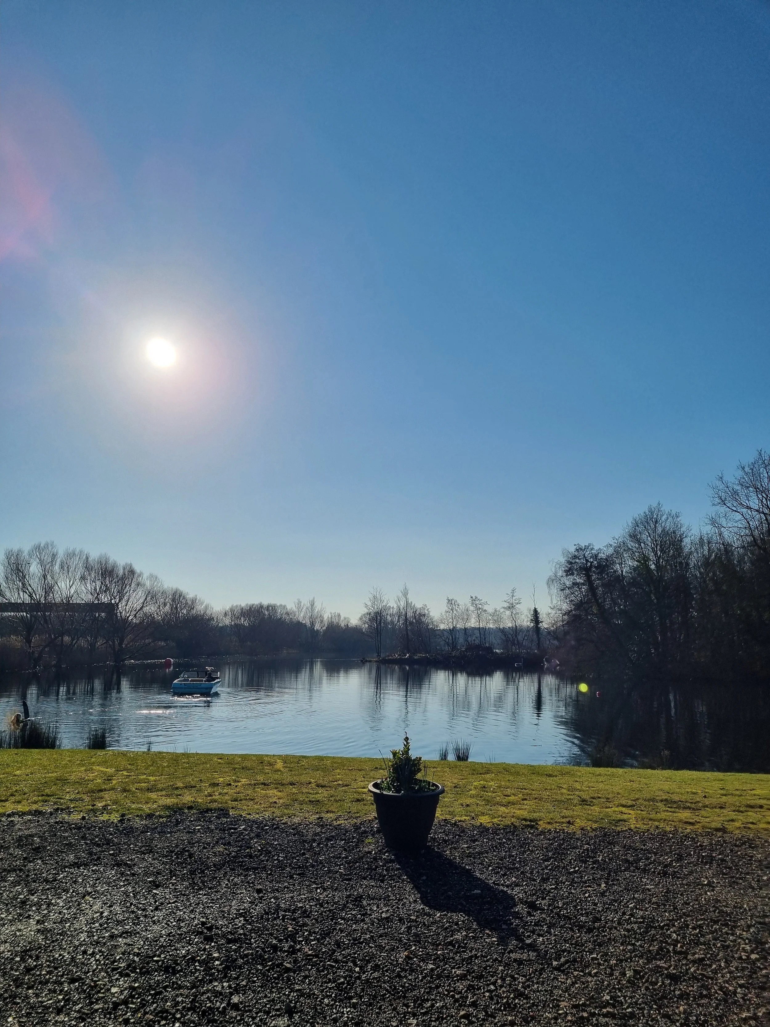 A calm river with a boat floating on it, surrounded by leafless trees, under a clear blue sky with a bright sun.