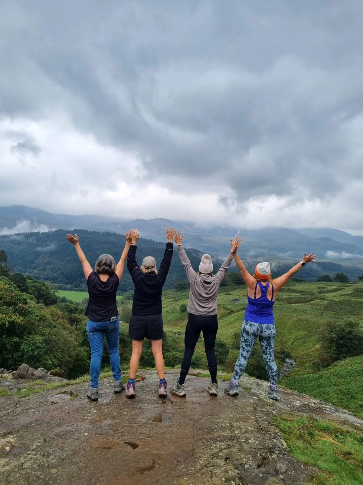 Four women with their backs to the camera, standing with arms raised, on a rocky hilltop overlooking lush green valleys and mountains under a cloudy sky.