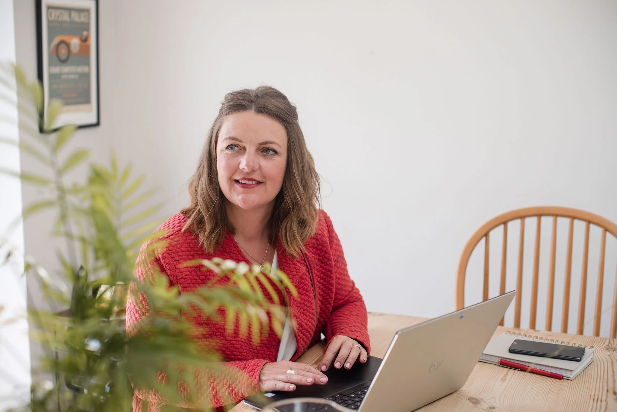 A woman with shoulder-length brown hair sitting at a wooden table with a laptop, notepad, and smartphone, working or having a meeting in a bright room with white walls and a wooden chair. A green plant and a framed poster are also visible.