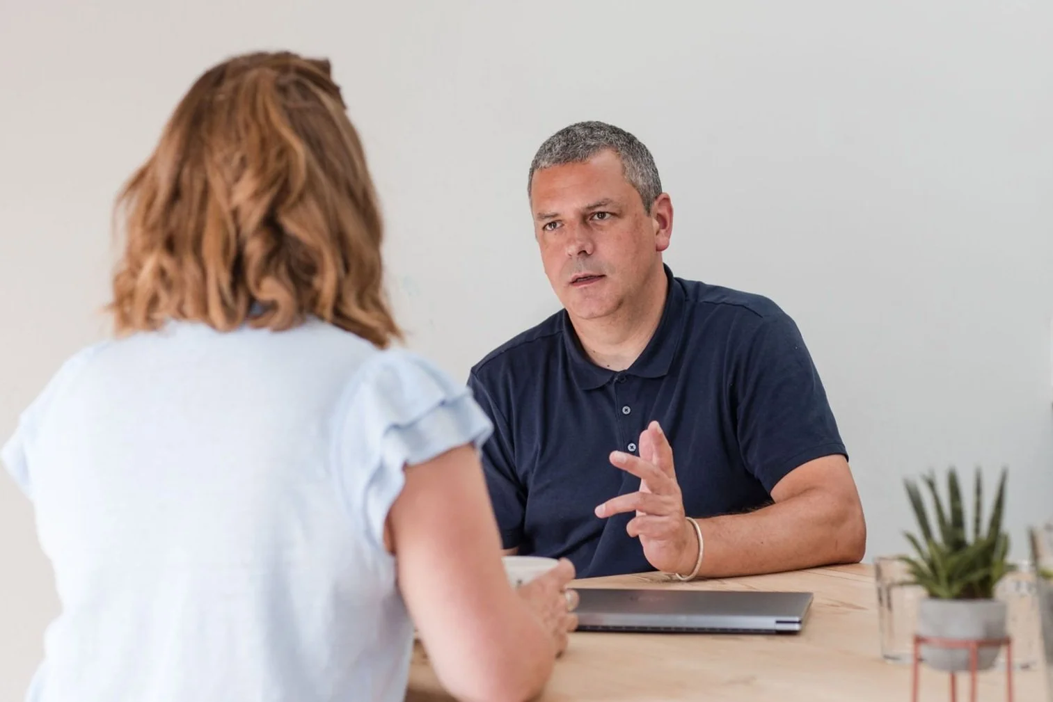 A man with short gray hair and a woman with shoulder-length red hair having a serious conversation at a wooden table, with a laptop and small potted plant in the background.