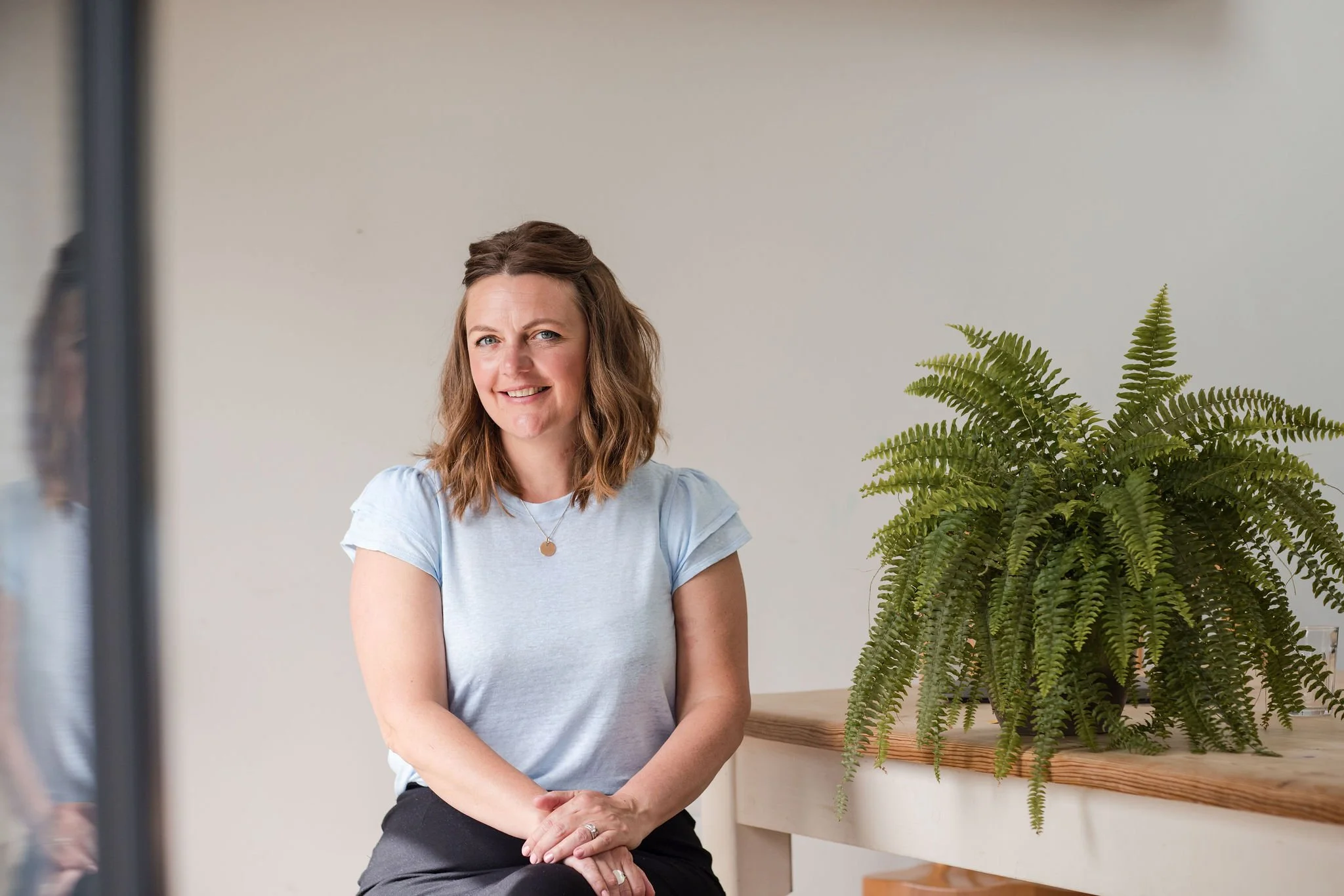 A woman with brown wavy hair and blue eyes sitting indoors next to a large green fern plant on a wooden bench, with a neutral background.