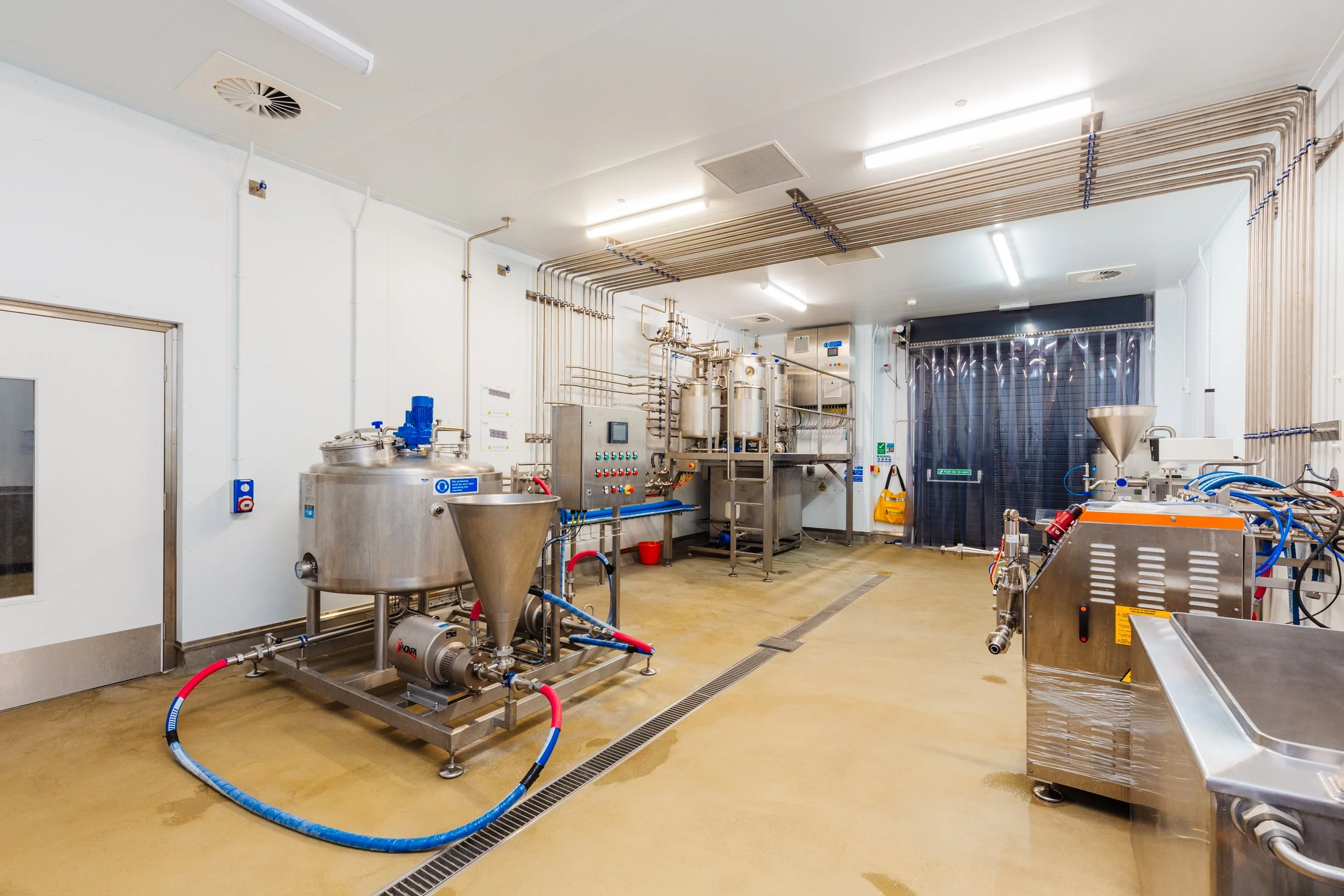 Two workers in white lab coats and knee-high boots (PPE) are operating stainless steel processing equipment in a clean industrial dairy facility, with control panels, pipes, and tanks involved in the production process.