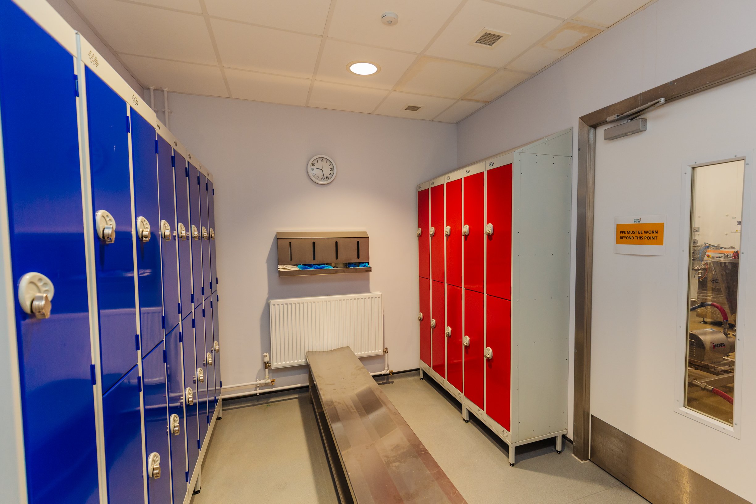 A locker room with two rows of lockers, blue on the left and red on the right. There is a clock on the wall showing 2:42, a bench, and a white wall with a radiator. An emergency exit door has a sign that reads 'PPE must be worn beyond this point' and a glass window showing the adjoining room.