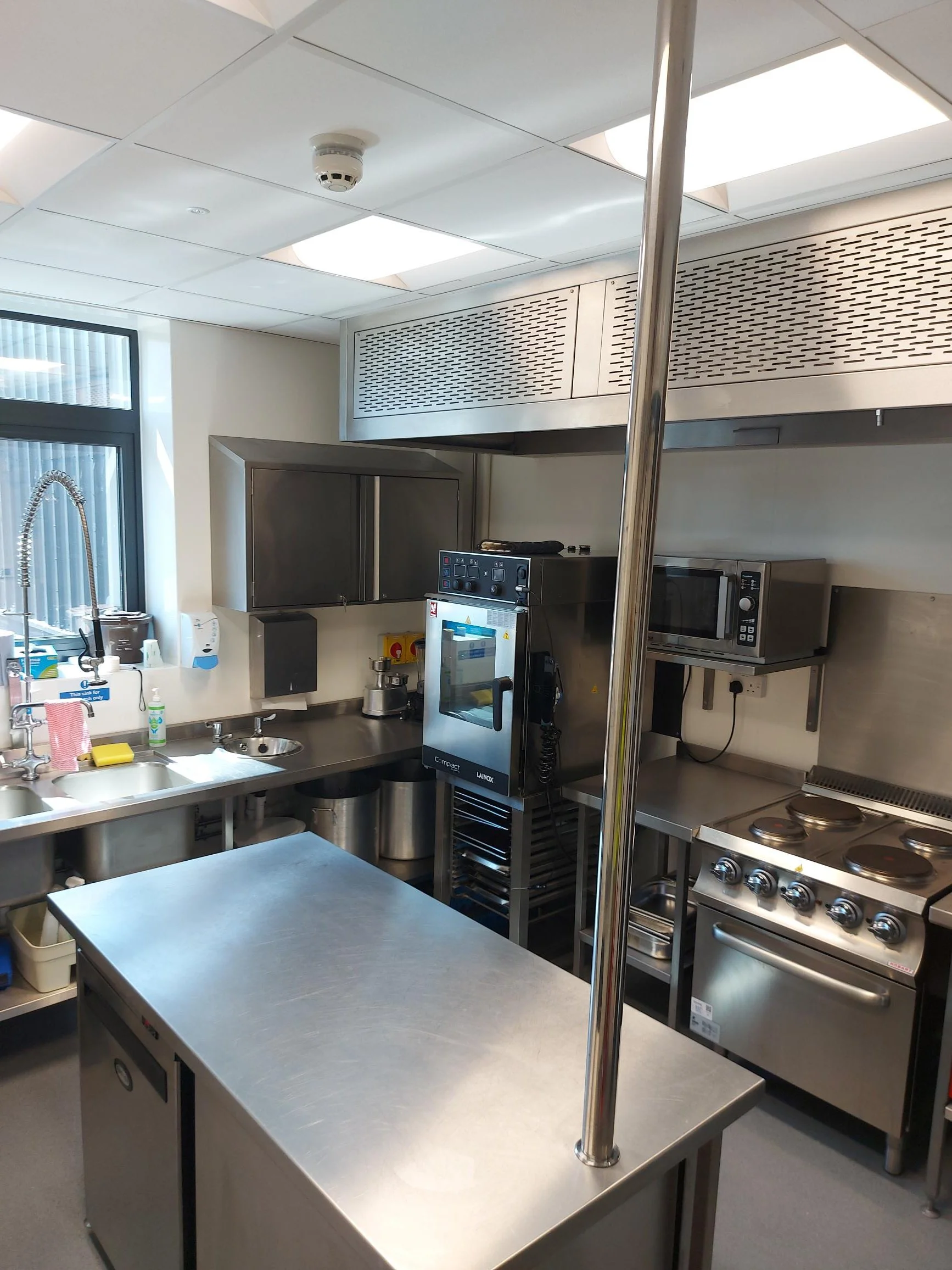 A commercial kitchen with stainless steel tables, appliances including an oven and microwave, a sink, and storage cabinets, illuminated by ceiling lights.