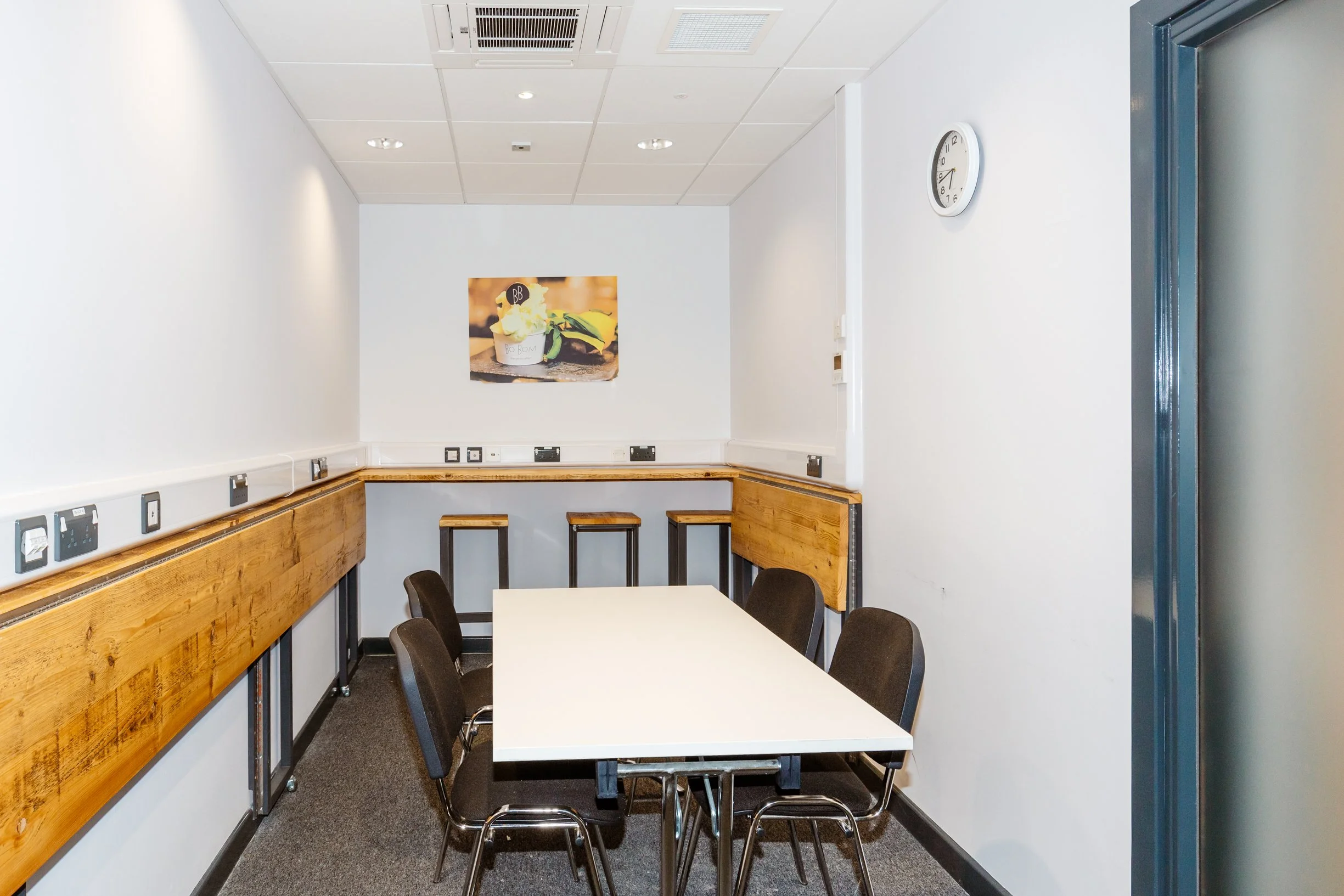 A conference or meeting room with a wall-mounted large flat-screen TV, wooden workbenches on the left and right with small matching stools, multiple electrical outlets and cords, and a white control panel on the right wall.