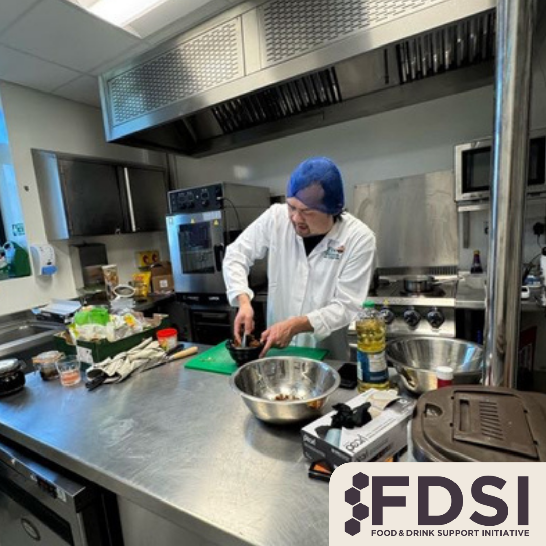A person in a white chef's coat and blue hairnet preparing food in a commercial kitchen, surrounded by various cooking utensils and ingredients. FDSI logo in the bottom right corner.
