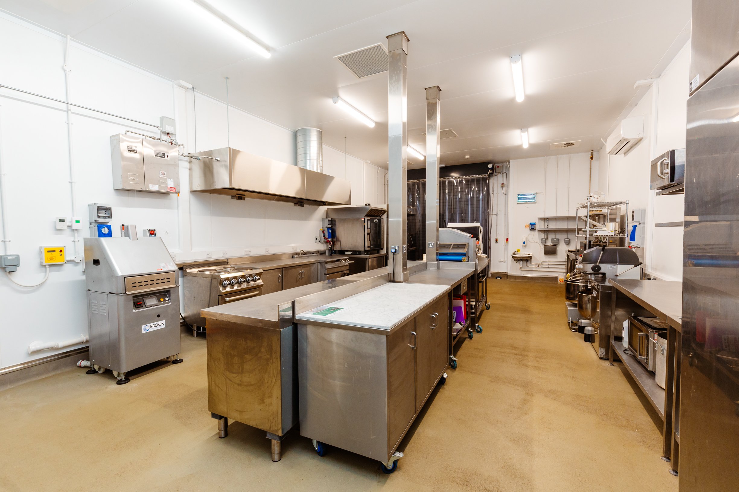 A woman in a lab coat and blue hairnet (PPE) smiling while operating a pasta dough sheet machine in a food processing facility.
