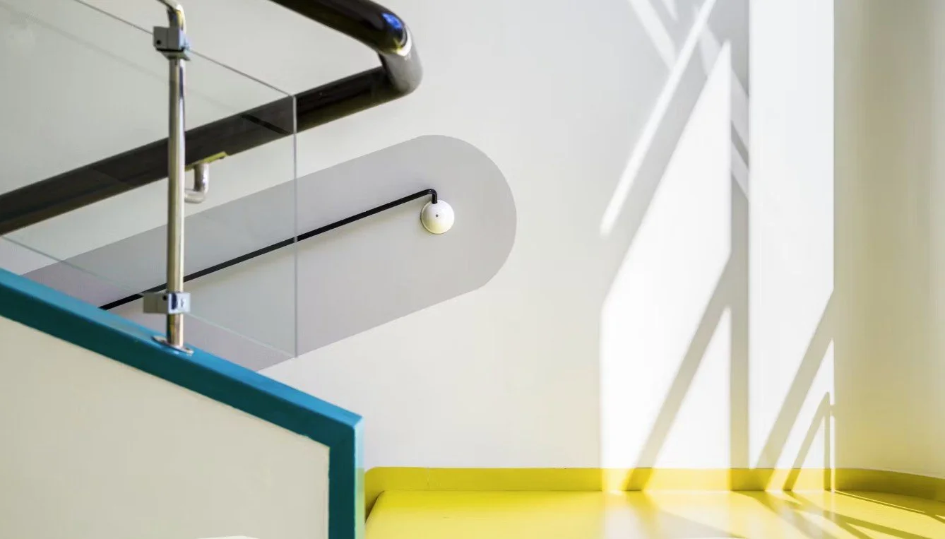 Modern stairwell with glass railing, yellow floor, sculptural shadows, and minimalist wall lighting.