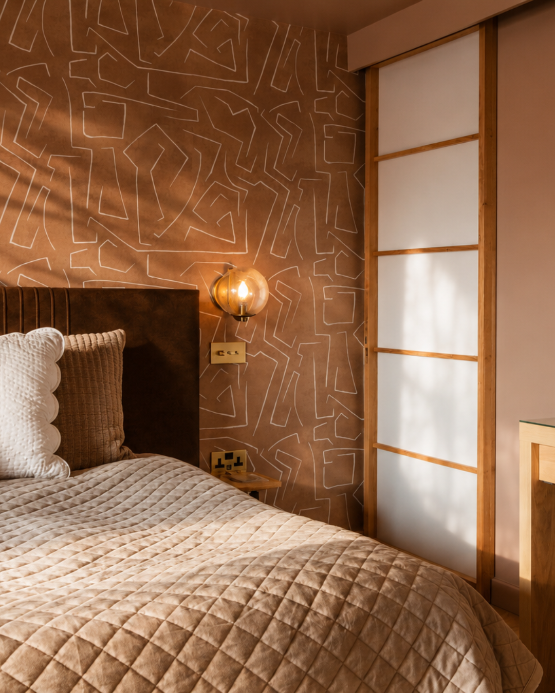 Warm evening light enhances the layered materials and textures within this bedroom in Great Shelford, Cambridge. Patterned wall coverings, natural timber finishes and soft upholstered elements work together to create a calm and characterful space tha