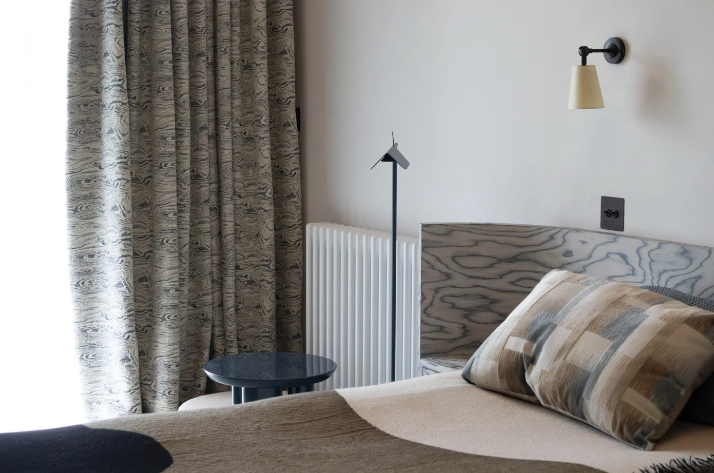 Bedroom detail in a Cambridge Water Lane new‑build renovation, featuring a bespoke wood‑veneer headboard, custom fabric‑matched curtains with a tailored pelmet, and layered neutral bedding in a calm contemporary palette