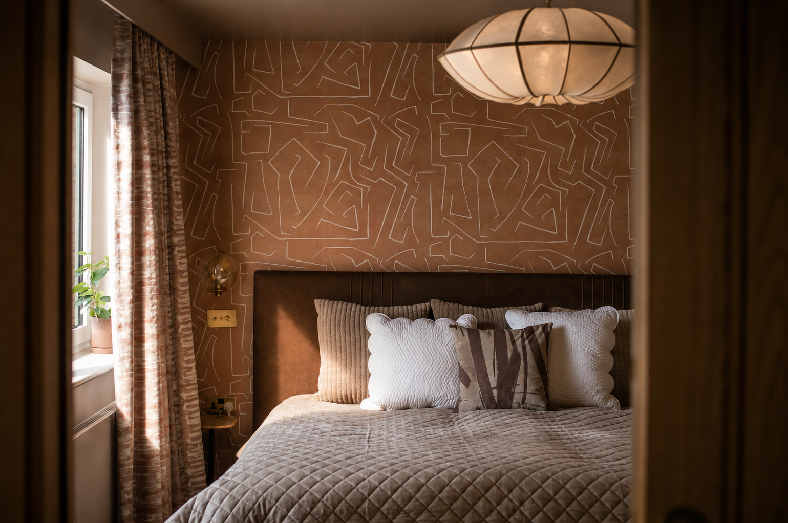 A warm and layered bedroom designed for a family home in Great Shelford, Cambridge. Textured wall coverings, rich timber tones and soft natural fabrics create a calm and inviting retreat, while carefully considered lighting adds depth and atmosphere 